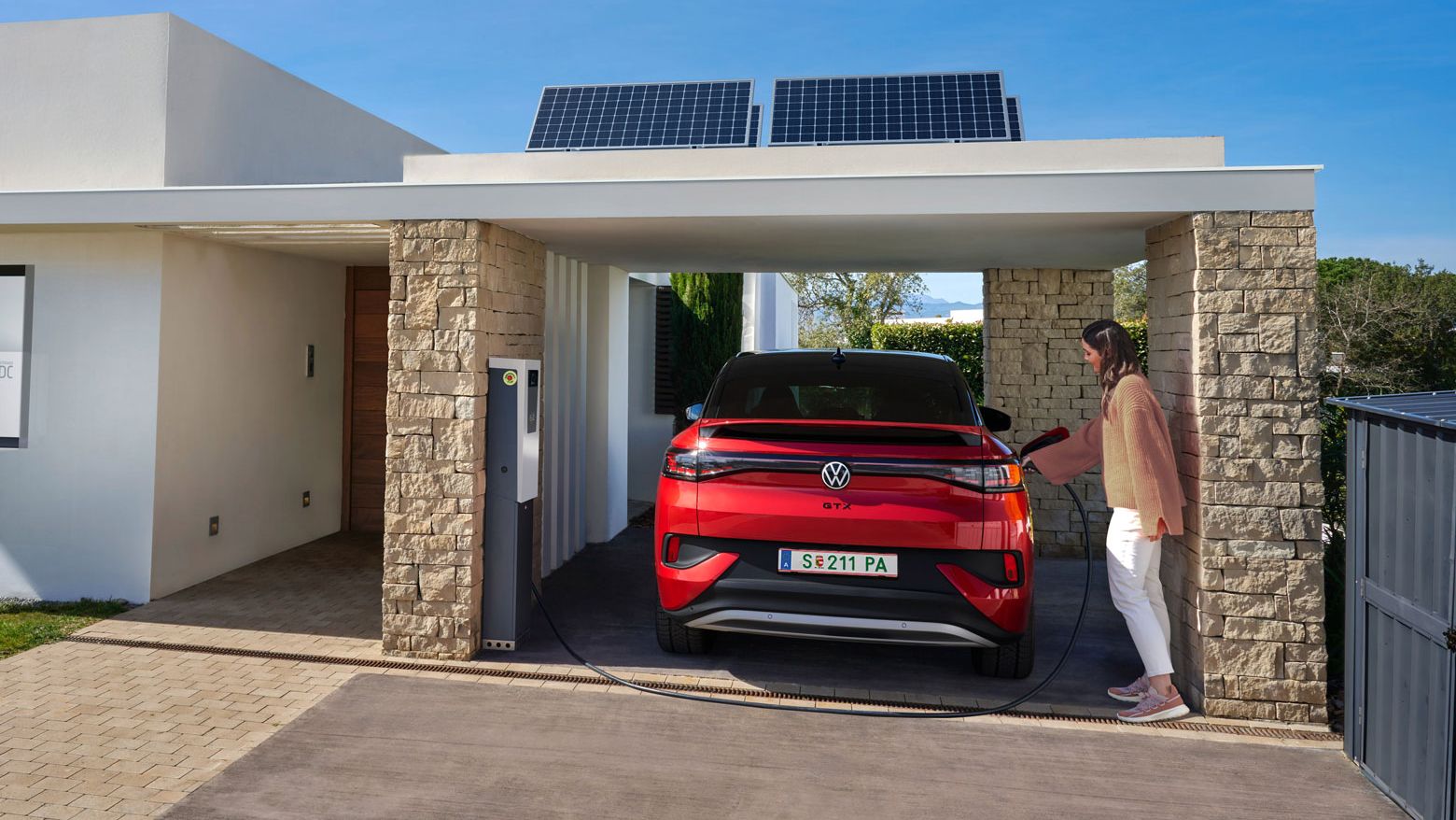 VW ID.5 GTX zaparkovaný pod prístreškom pri dome je napojený na domácu nabíjaciu stanicu a žena pripája k vozidlu nabíjací kábel. 