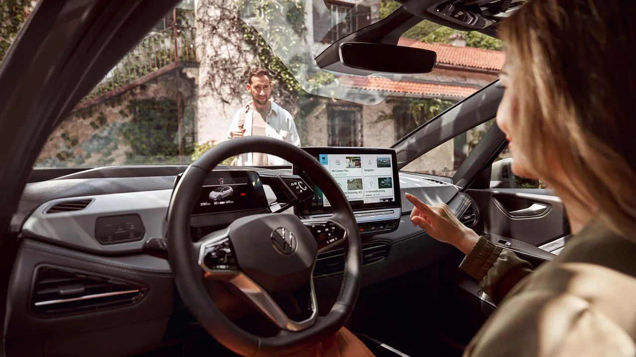 Interiér moderného vozidla s digitálnymi displejmi a dotykovým ovládaním. Žena sedí na sedadle vodiča a ovláda funkcie systému infotainment. Von pred vozidlom stojí muž a pozerá sa na ženu vo vozidle.