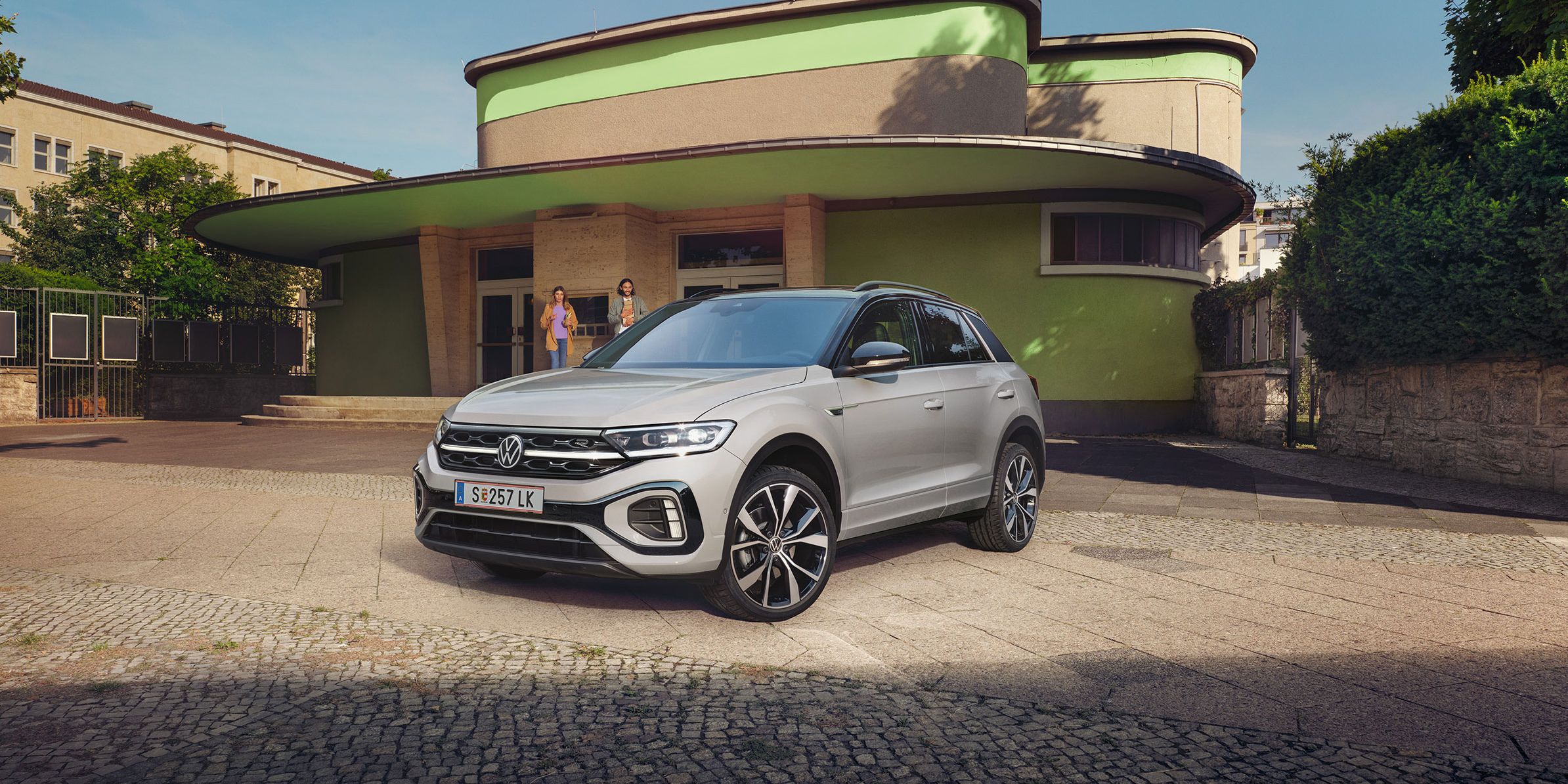 Strieborné SUV Volkswagen T-Roc je zaparkované na ulici pred budovou so svetlohnedou, zelenou fasádou a zaoblenými tvarmi. Budova je obklopená zeleňou. 