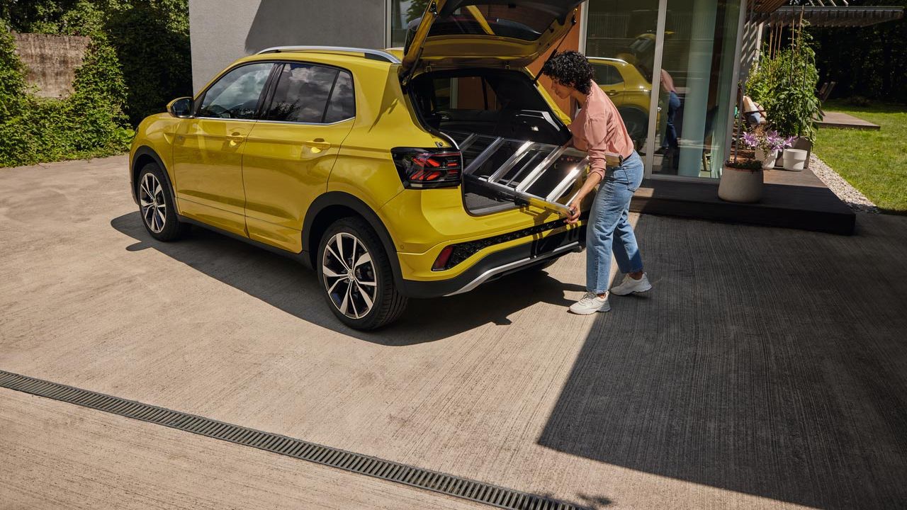Mladá žena vkladá rebrík do batožinového priestoru žltého VW T-Cross. Vozidlo je zaparkované pred moderným rodinným domom s peknou záhradou s množstvom zelene.