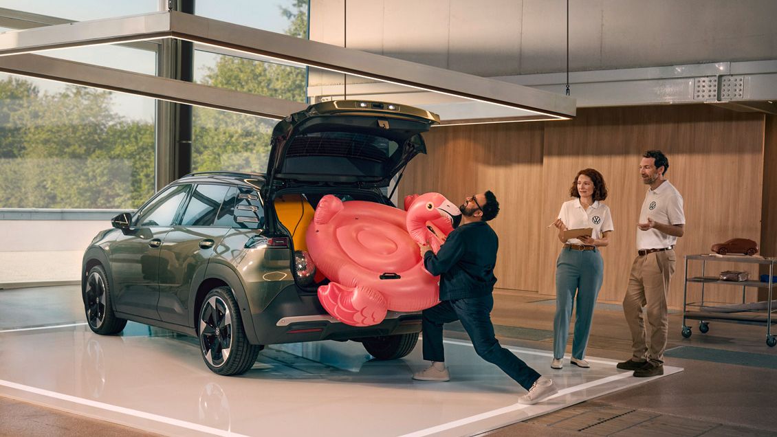 Ein Mann drückt einen aufblasbaren Flamingo Muž vkladá nafukovacieho plameniaka do otvoreného batožinového priestoru vozidla VW ID. Cross. Vedľa neho stoja dve osoby a so záujmom ho pozorujú.in den offenen Kofferraum des VW ID. Cross. Zwei Personen stehen daneben und gucken ihm amüsiert zu.