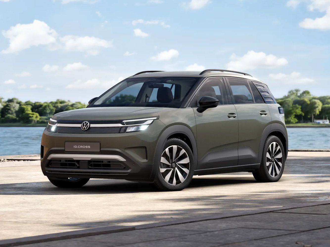 Olivovo-zelený VW ID. Cross Life s moderným, hranatým dizajnom stojí na promenáde pri vode pred jazerom. V pozadí je vidieť vodu, stromy a modrú oblohu s roztrúsenými oblakmi.