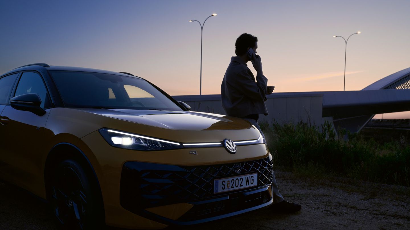 Muž sa opiera o vozidlo VW T-Roc R-Line so zapnutými svetlami a osvetleným logom.