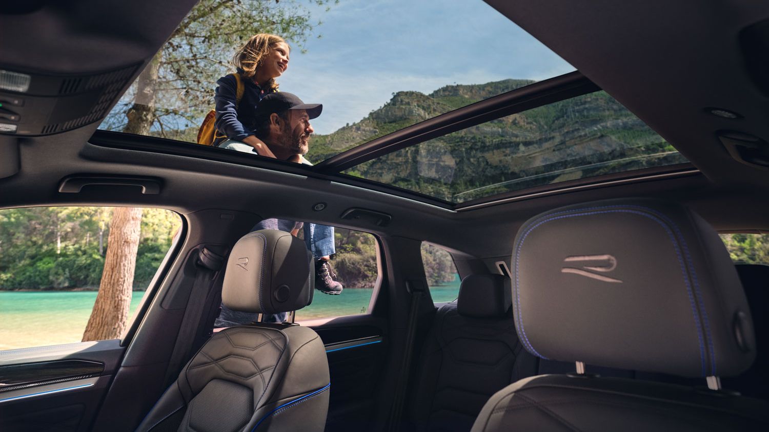 Pohľad cez panoramatickú strechu VW Touareg R eHybrid na okolie a muža s dcérou na pleciach. Čiastočne vidieť aj čierne predné sedadlá s logom R.