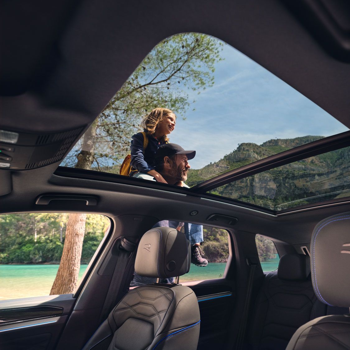 Pohľad cez panoramatickú strechu VW Touareg R eHybrid na okolie a muža s dcérou na pleciach. Čiastočne vidieť aj čierne predné sedadlá s logom R.