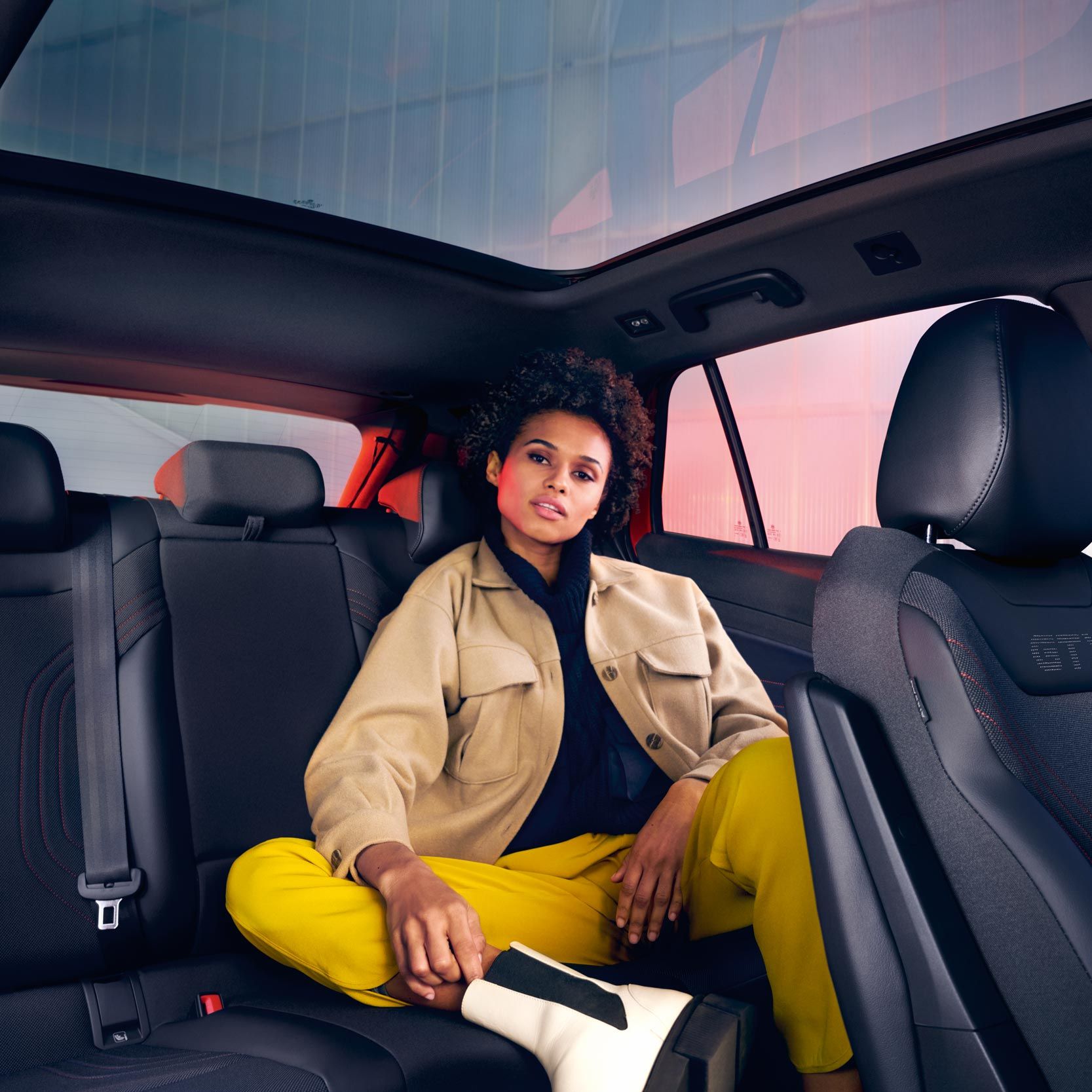 Pohľad do interiéru vozidla VW ID.4 GTX, žena sedí na zadnom sedadle otočená dopredu a pozerá sa priamo do fotoaparátu. Cez okná je vidieť modernú budovu.