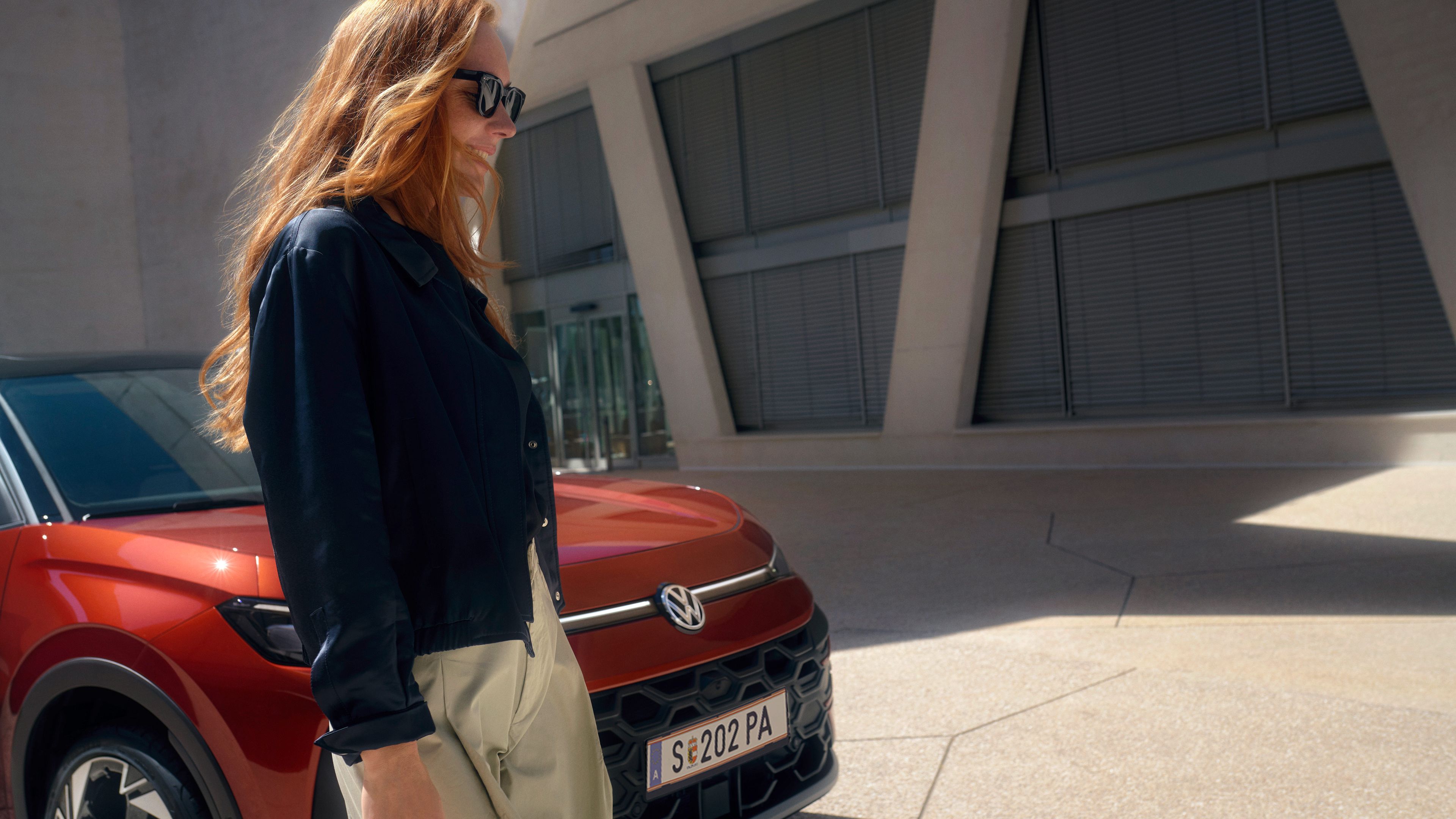 Žena v slnečných okuliaroch stojí vedľa červeného VW TRoc Style pred budovou.