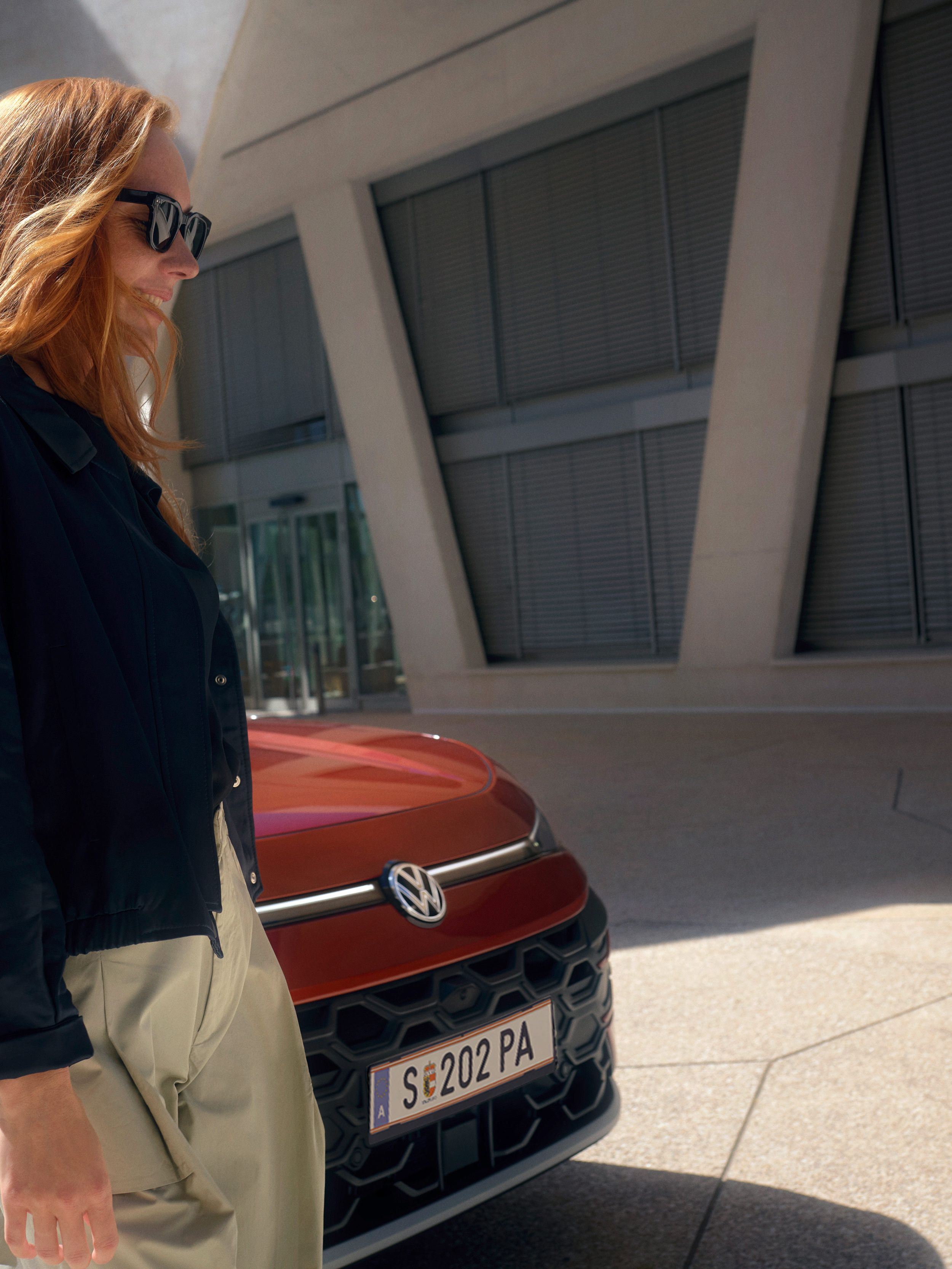 Žena v slnečných okuliaroch stojí vedľa červeného VW TRoc Style pred budovou.