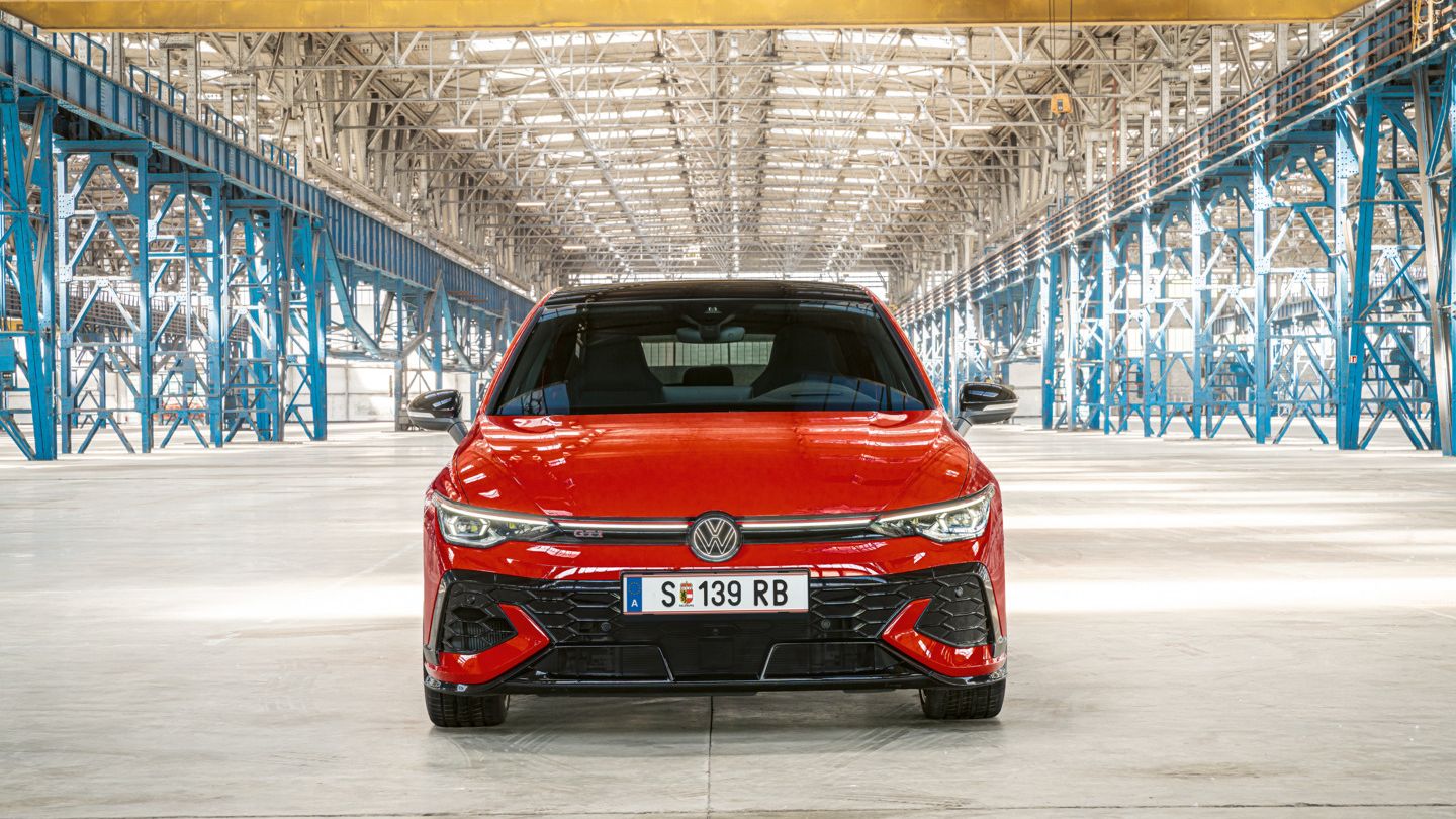 Červený Golf GTI je zaparkovaný vo veľkom, jasne osvetlenom sklade. Auto je zobrazené spredu. V pozadí sú viditeľné kovové konštrukcie a stĺpy.