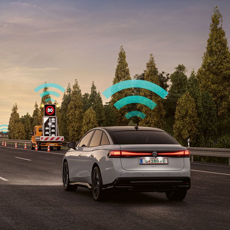VW Varovanie pred nebezpečenstvom Car2X zobrazené ako modrý ikona WiFi nad vozidlami, ktoré jazdia po diaľnici. Diaľnicu lemuje zeleň a stromy. Atmosféru dotvára modrá, čiastočne zatiahnutá obloha pri západe slnka.