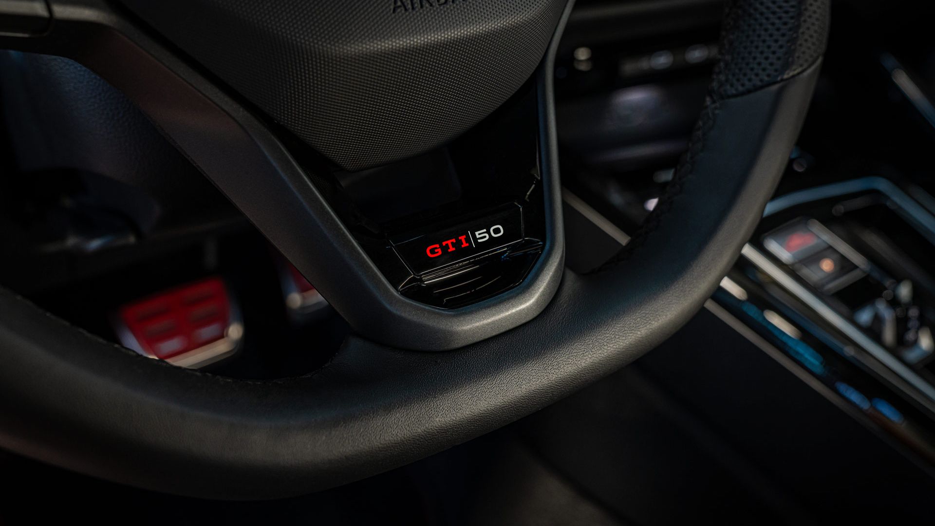 Detailný záber na volant s nápisom „GTI 50“ v aute. Čiastočne viditeľné sú aj ovládacie tlačidlá a konzola za nimi. Oblasť airbagu je označená slovom „AIRBAG“.