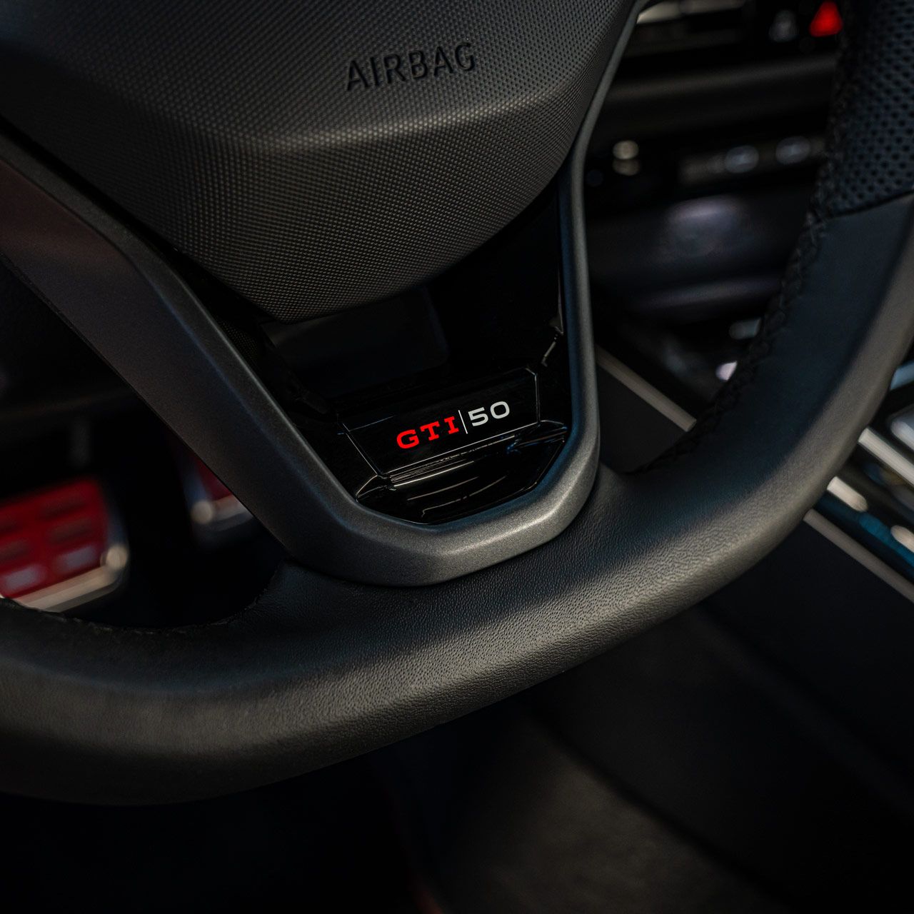 Detailný záber na volant s nápisom „GTI 50“ v aute. Čiastočne viditeľné sú aj ovládacie tlačidlá a konzola za nimi. Oblasť airbagu je označená slovom „AIRBAG“.