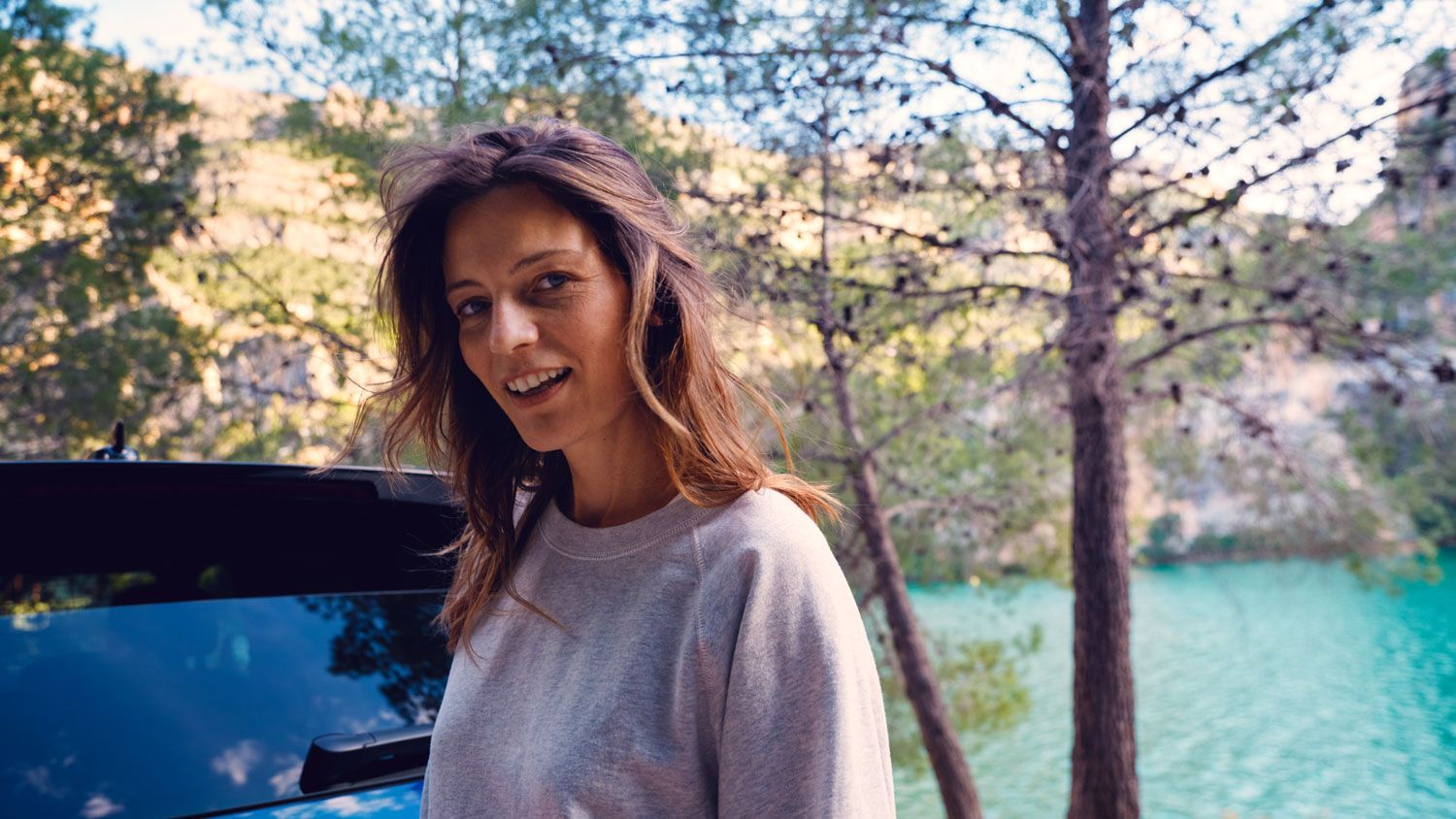 Usmiatá žena stojí pred vozidlom VW Touareg R eHybrid, ktorý stojí pri rieke v horskom prostredí.
