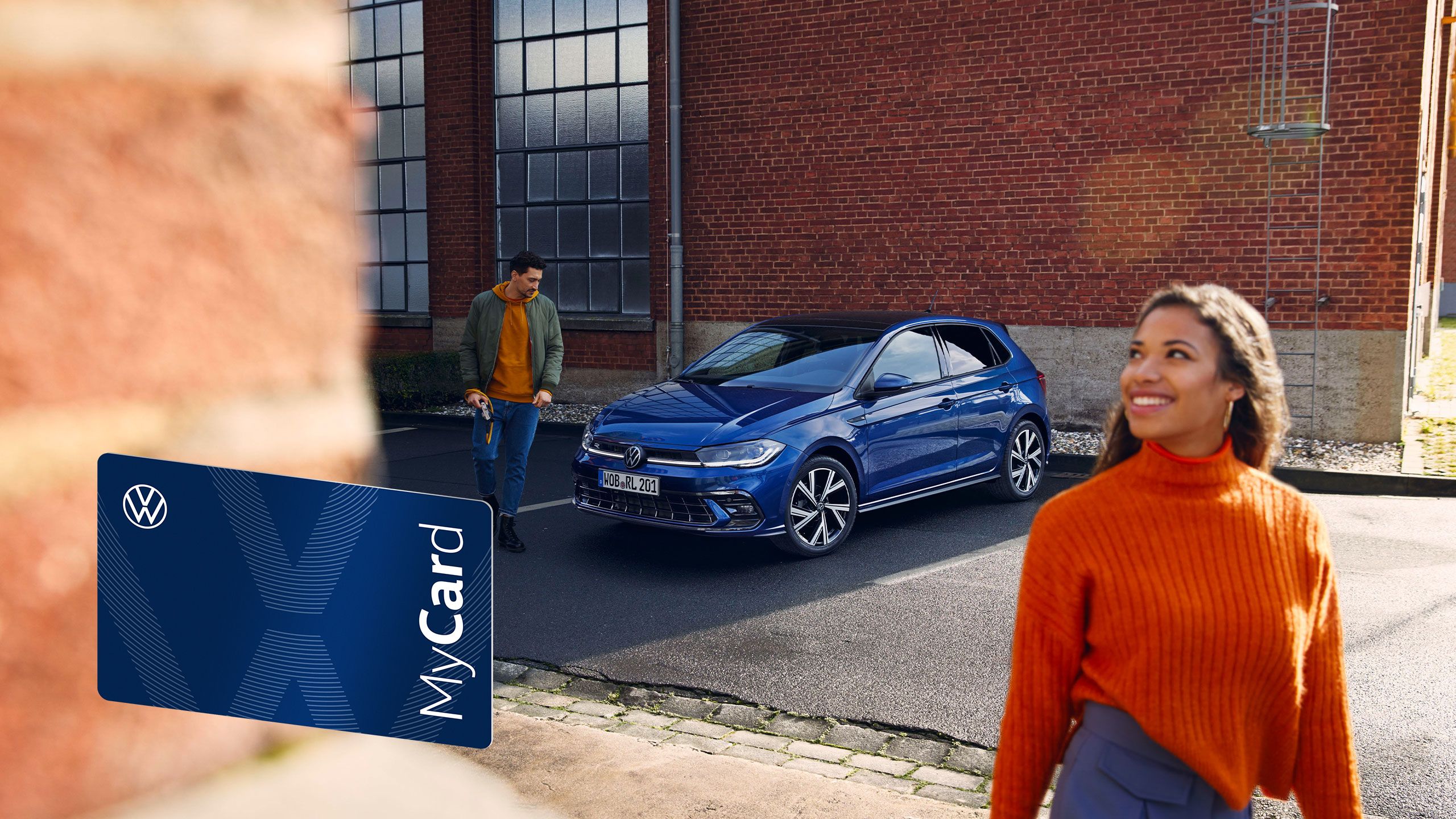 Modrý Golf zaparkovaný pri tehlovej industriálnej budove, pri vozidle stojí muž a pred vozidlom žena. 