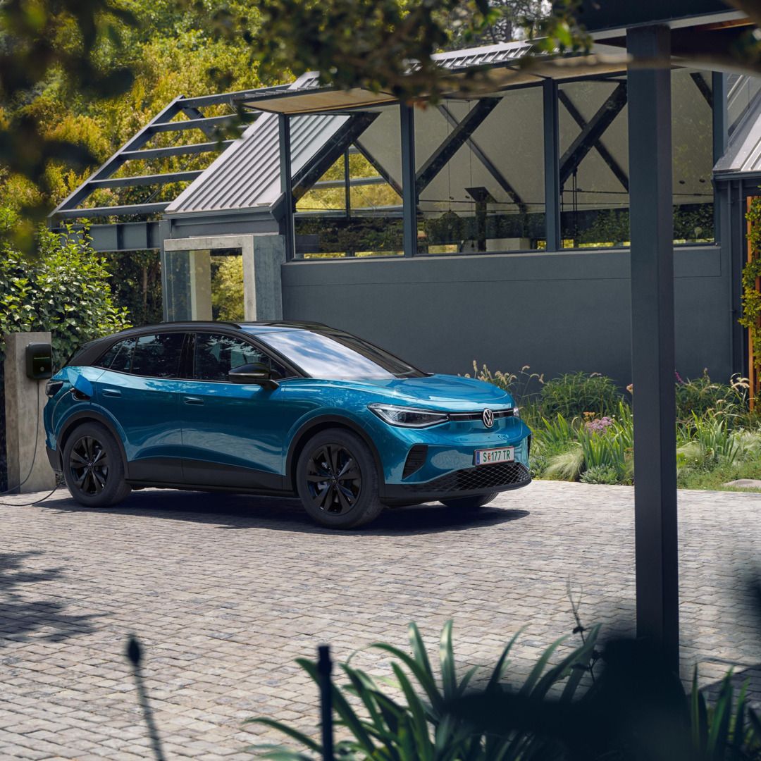 Modrý VW ID.4 s čiernou strechou parkuje vedľa moderného domu a nabíja sa na nabíjacej stanici. 