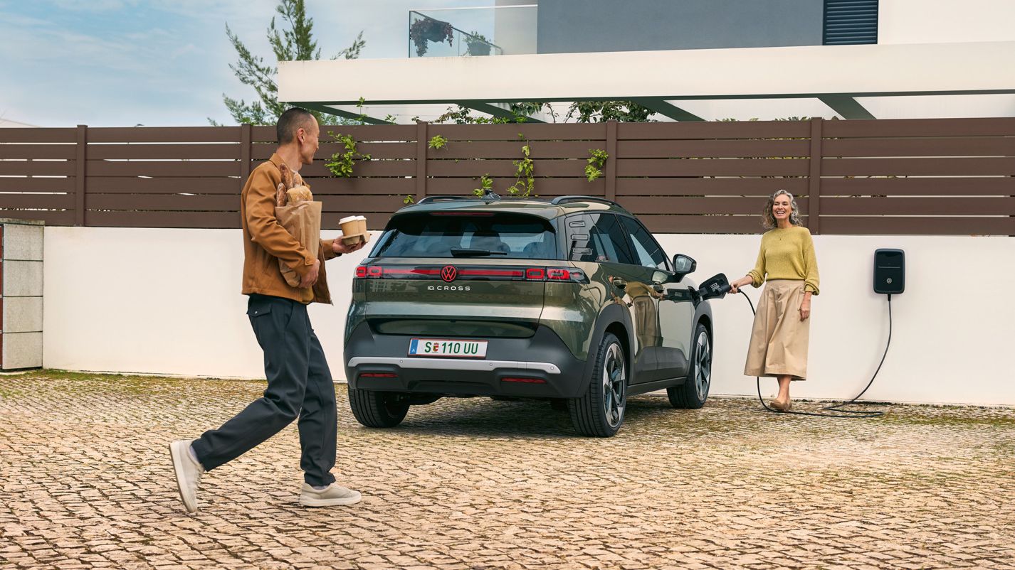 Žena zapája nabíjací kábel zo zásuvky do zeleného VW ID. Cross, muž, k nej pristúpi s kávou so sebou v ruke a pečivom.