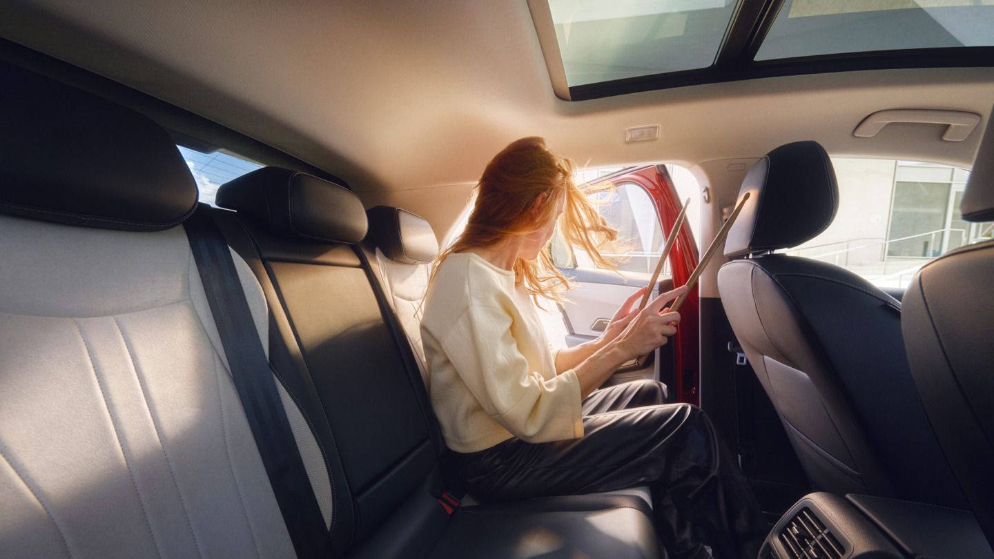 Žena s dlhými červenými vlasmi sedí na zadnom sedadle vozidla T-Roc. Pozerá sa von cez otvorené dvere a drží paličky od bubna. Interiér vozidla je moderný a pohodlný.