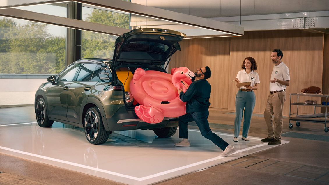 Ein Mann drückt einen aufblasbaren Flamingo Muž vkladá nafukovacieho plameniaka do otvoreného batožinového priestoru vozidla VW ID. Cross. Vedľa neho stoja dve osoby a so záujmom ho pozorujú.in den offenen Kofferraum des VW ID. Cross. Zwei Personen stehen daneben und gucken ihm amüsiert zu.