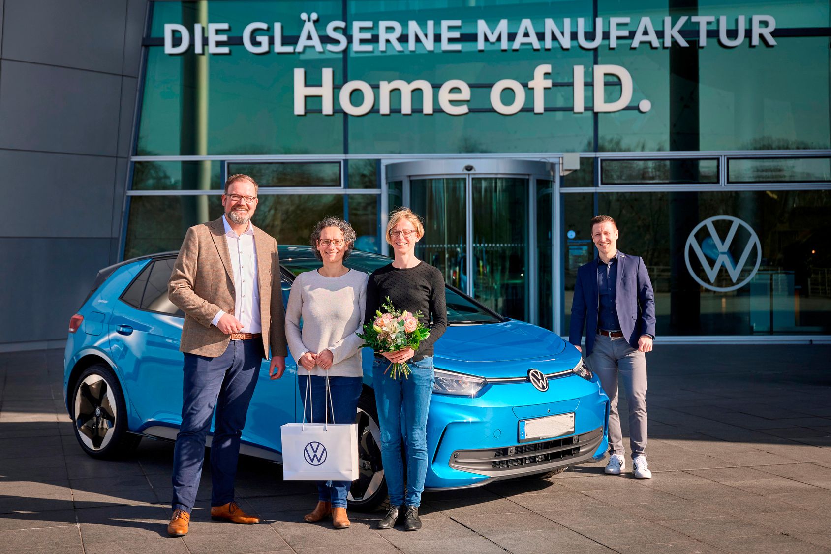 Skupina ľudí stojí pred budovou s nápisom "DIE GLÄSERNE MANUFAKTUR" a "Home of ID." Vedľa nich je zaparkované modré elektrické vozidlo Volkswagen ID.4. Jeden z ľudí drží darčekovú tašku s logom Volkswagen a iná osoba drží kyticu kvetov.