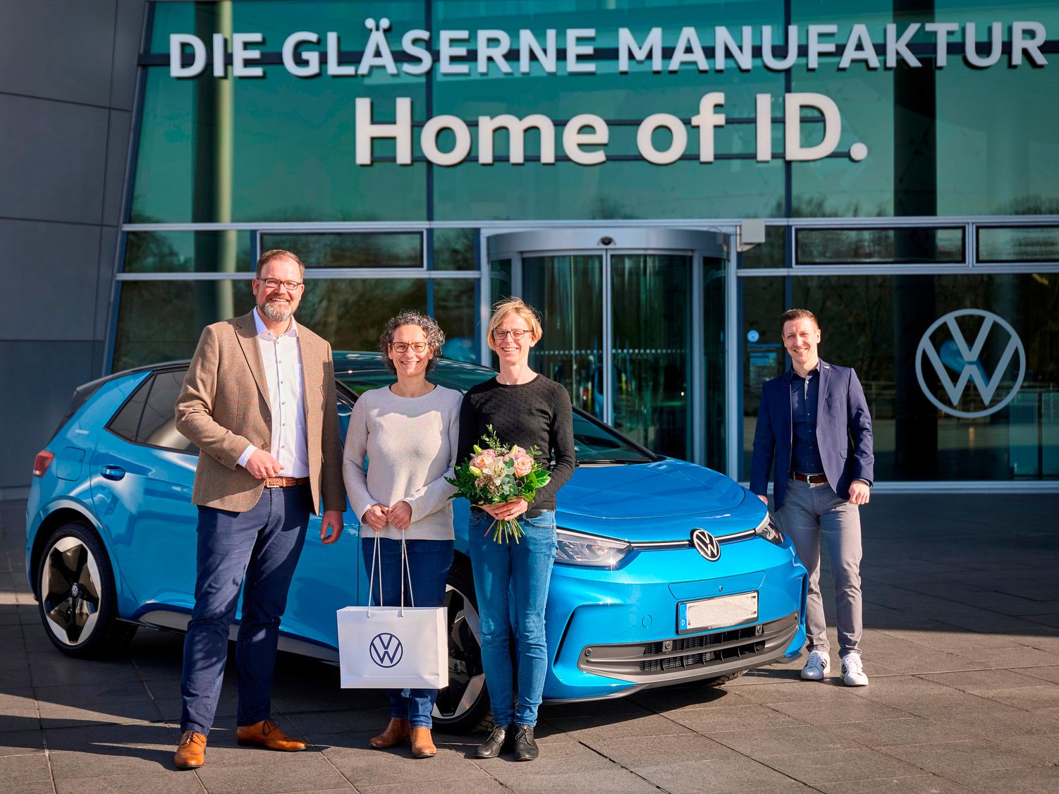 Skupina ľudí stojí pred budovou s nápisom "DIE GLÄSERNE MANUFAKTUR" a "Home of ID." Vedľa nich je zaparkované modré elektrické vozidlo Volkswagen ID.4. Jeden z ľudí drží darčekovú tašku s logom Volkswagen a iná osoba drží kyticu kvetov.