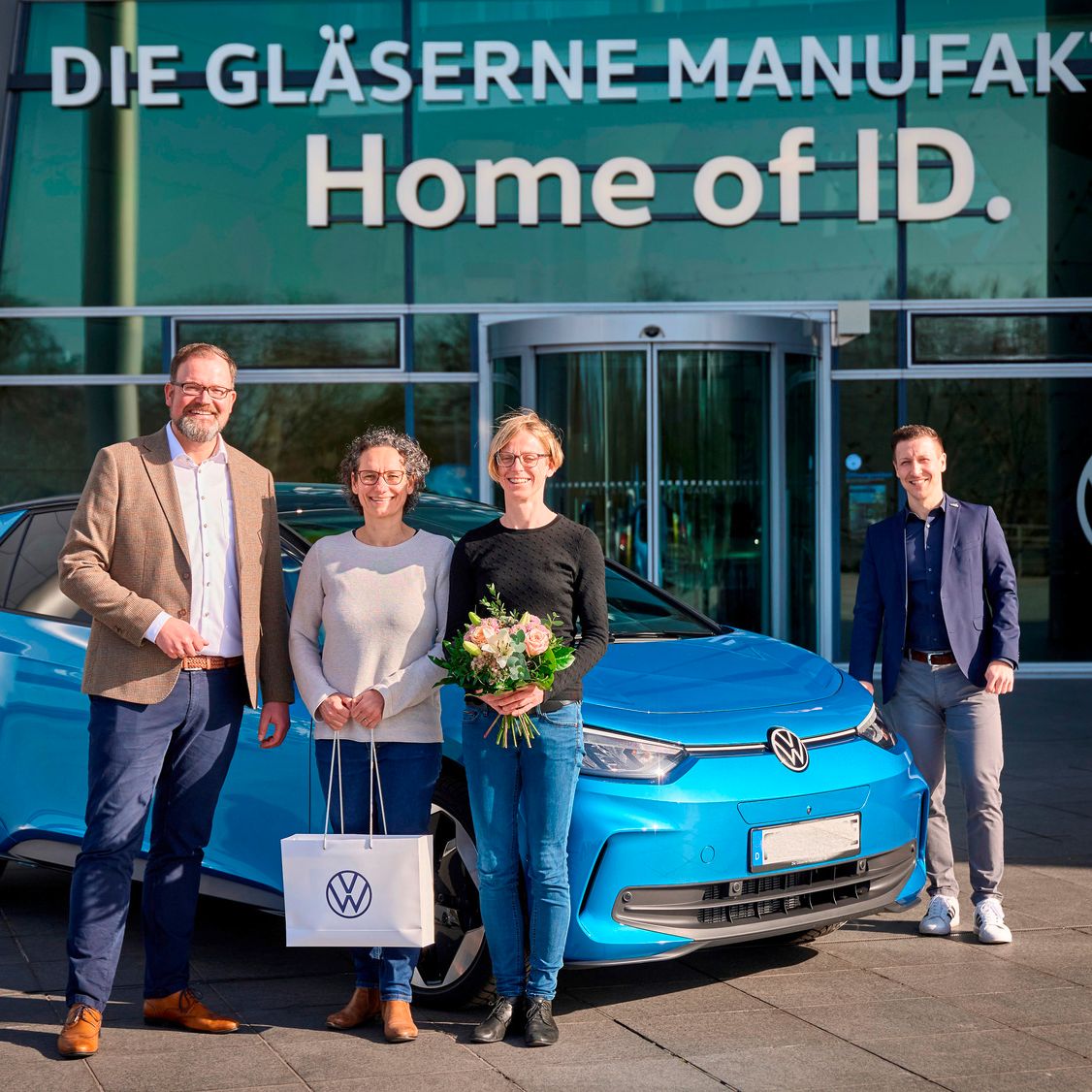 Skupina ľudí stojí pred budovou s nápisom "DIE GLÄSERNE MANUFAKTUR" a "Home of ID." Vedľa nich je zaparkované modré elektrické vozidlo Volkswagen ID.4. Jeden z ľudí drží darčekovú tašku s logom Volkswagen a iná osoba drží kyticu kvetov.