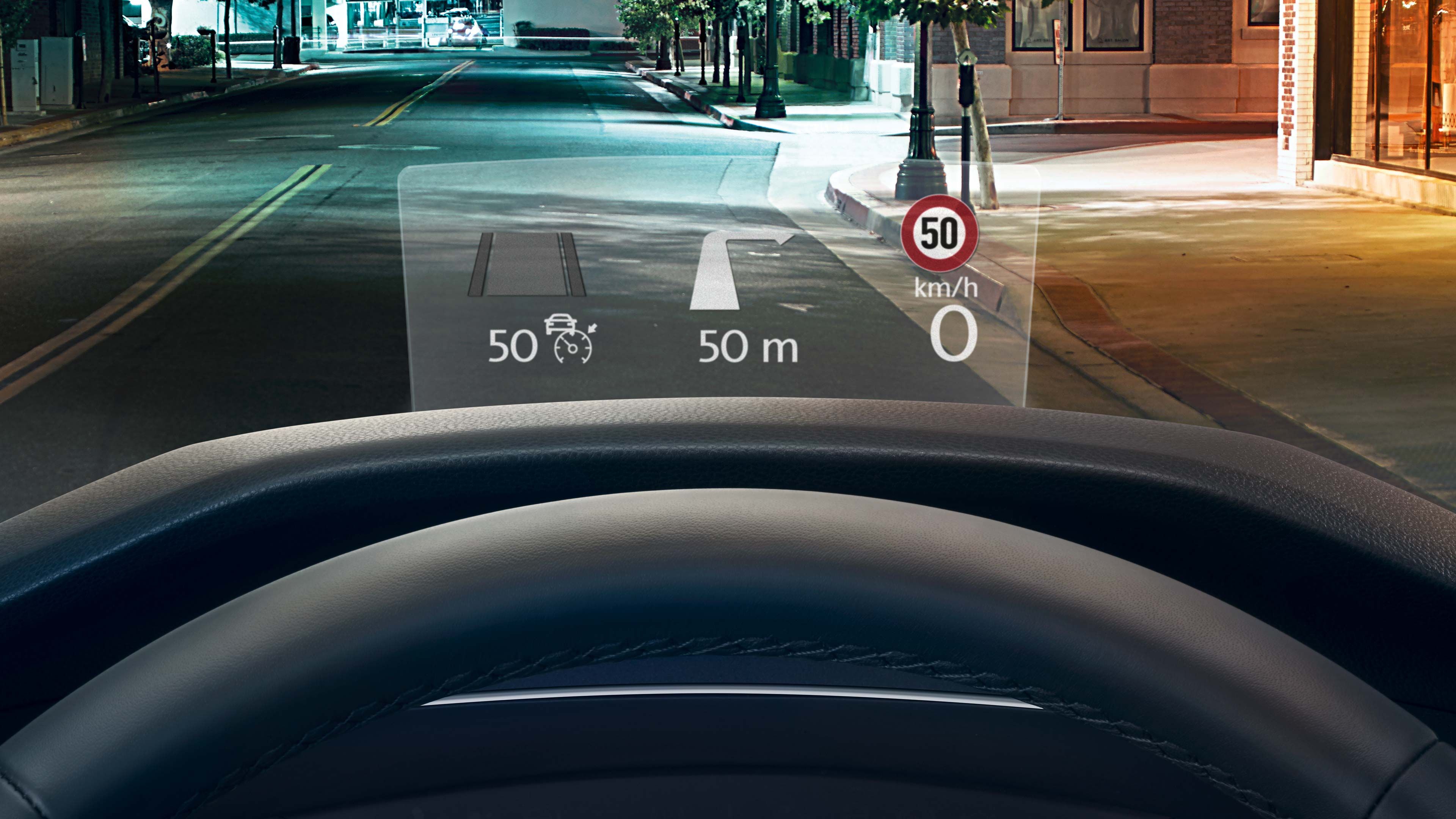 Head-up displej v automobile ukazuje informácie o aktuálnej rýchlosti, povolenom rýchlostnom limite a vzdialenosti. V displeji sú znázornené aj jazdné pruhy na vozovke. Von je noc a ulice sú osvetlené. Cez okno vidieť mesto.