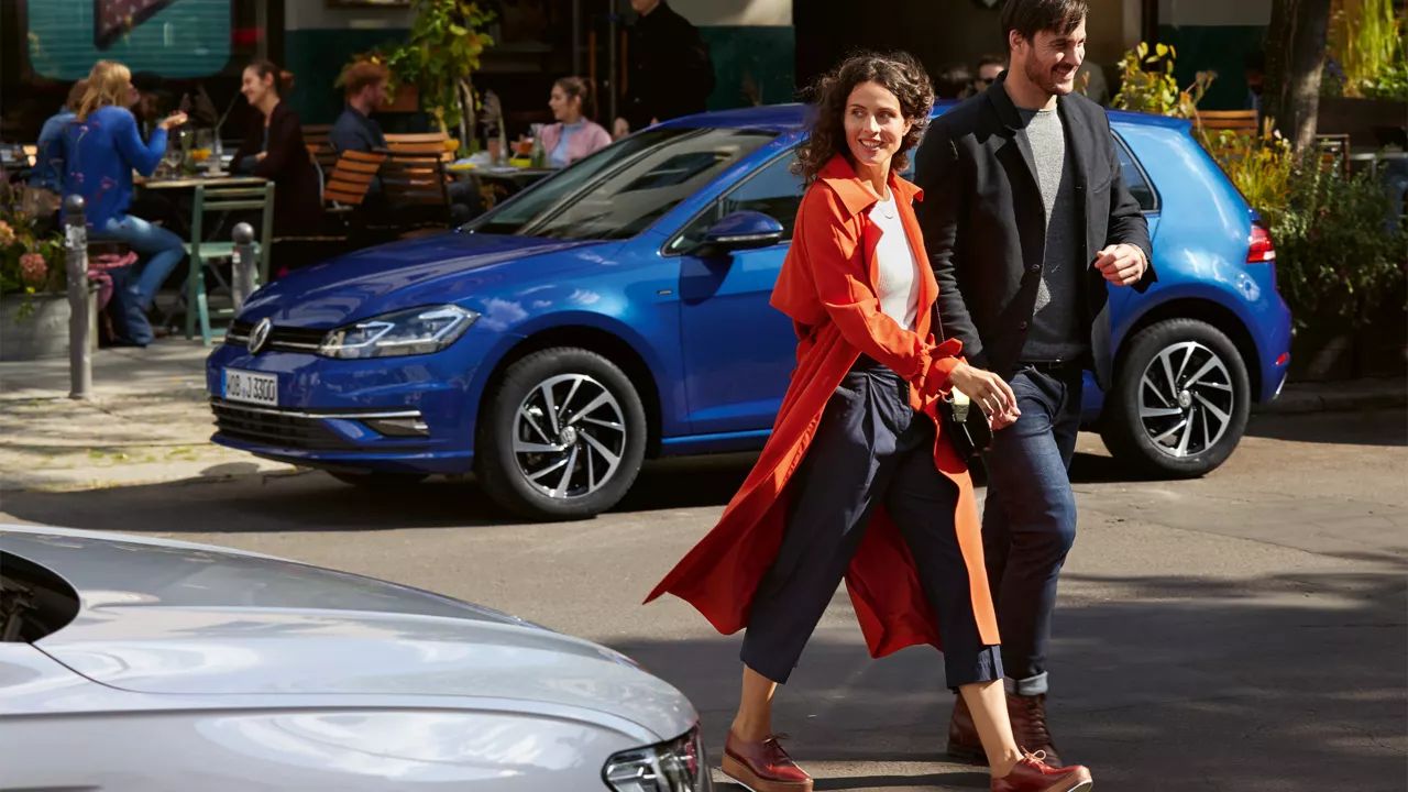 Žena a muž prechádzajú cez cestu. V pozadí stojí modrý VW Golf pred kaviarňou.