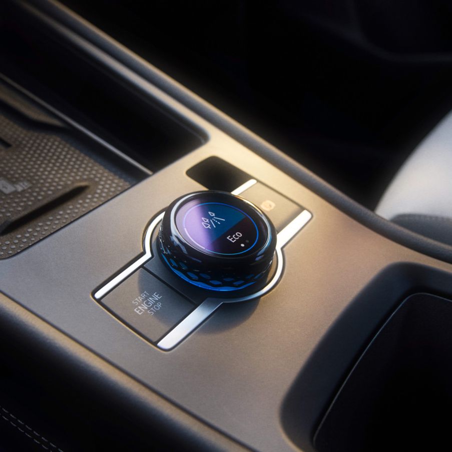 Detailný záber stredovej konzoly v modeli T-Roc so zameraním na prepínač jazdných režimov. Okrúhle otočné tlačidlo s LED ukazovateľmi na ovládanie rôznych funkcií vozidla. Viditeľné sú vysokokvalitné materiály a elegantný dizajn.
