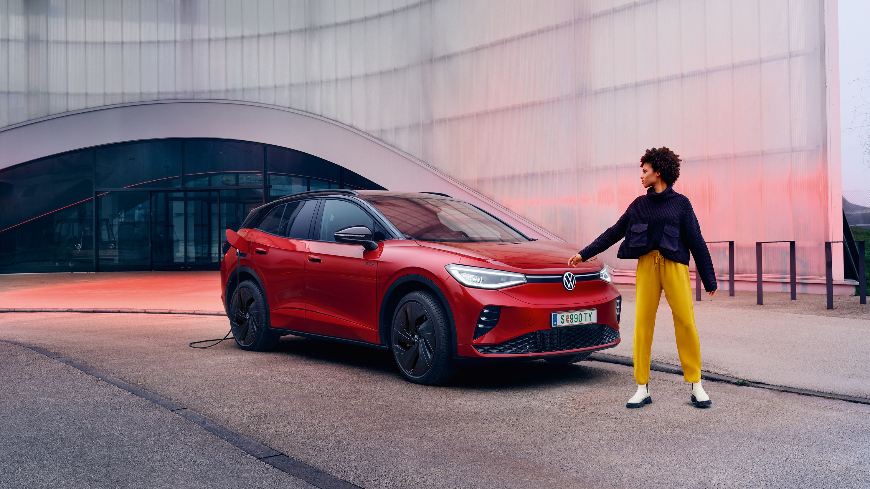 Žena v žltých nohaviciach a tmavej bunde stojí vedľa červeného SUV Volkswagen ID.4 GTX, ktoré je zaparkované pred modernou budovou. Na pozadí je viditeľná priehľadná fasáda modernej budovy.