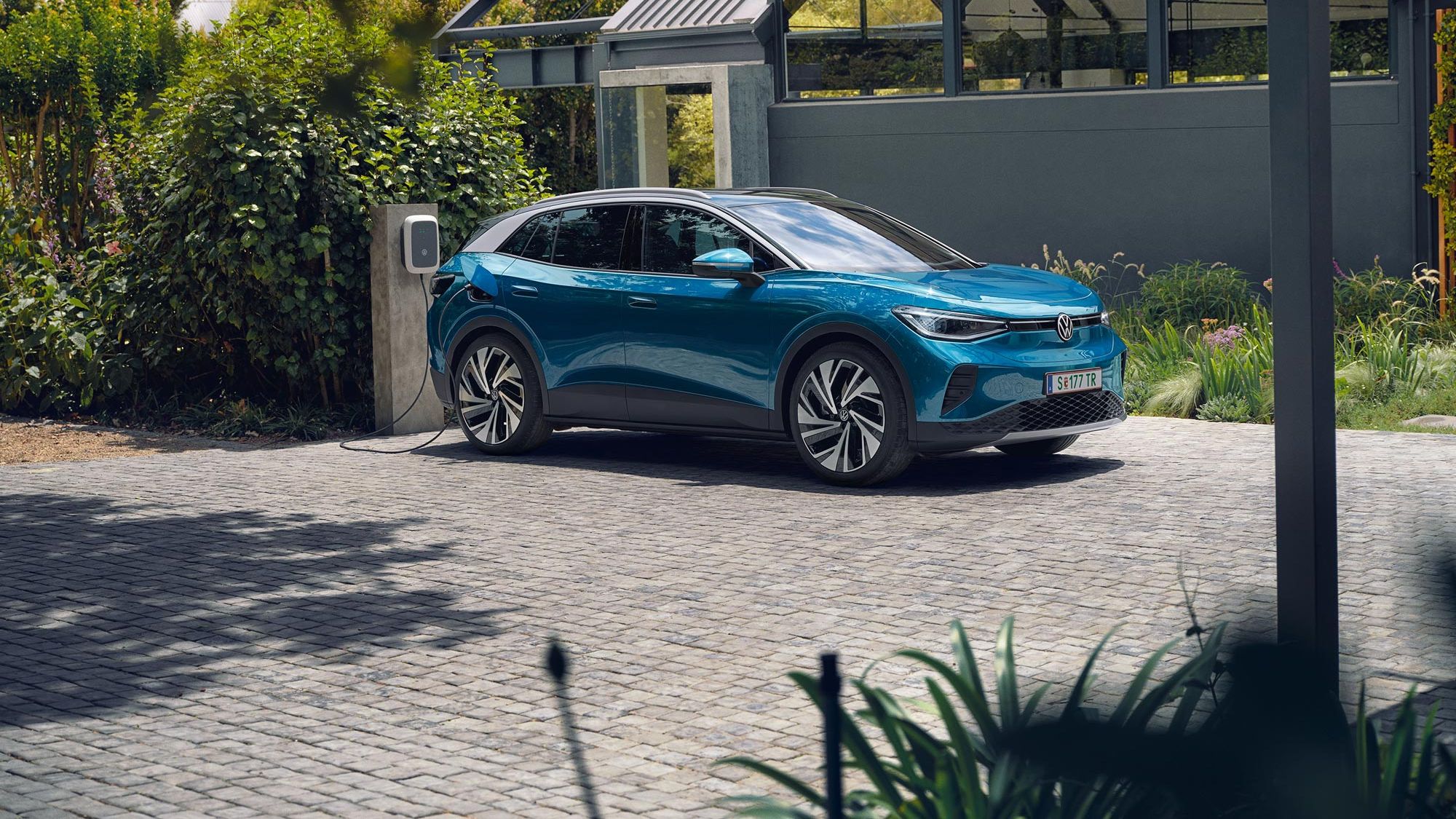 Modré elektrické SUV značky Volkswagen stojí na dláždenej ploche pri domácej nabíjacej stanici. V pozadí je zeleň a malá moderná budova.