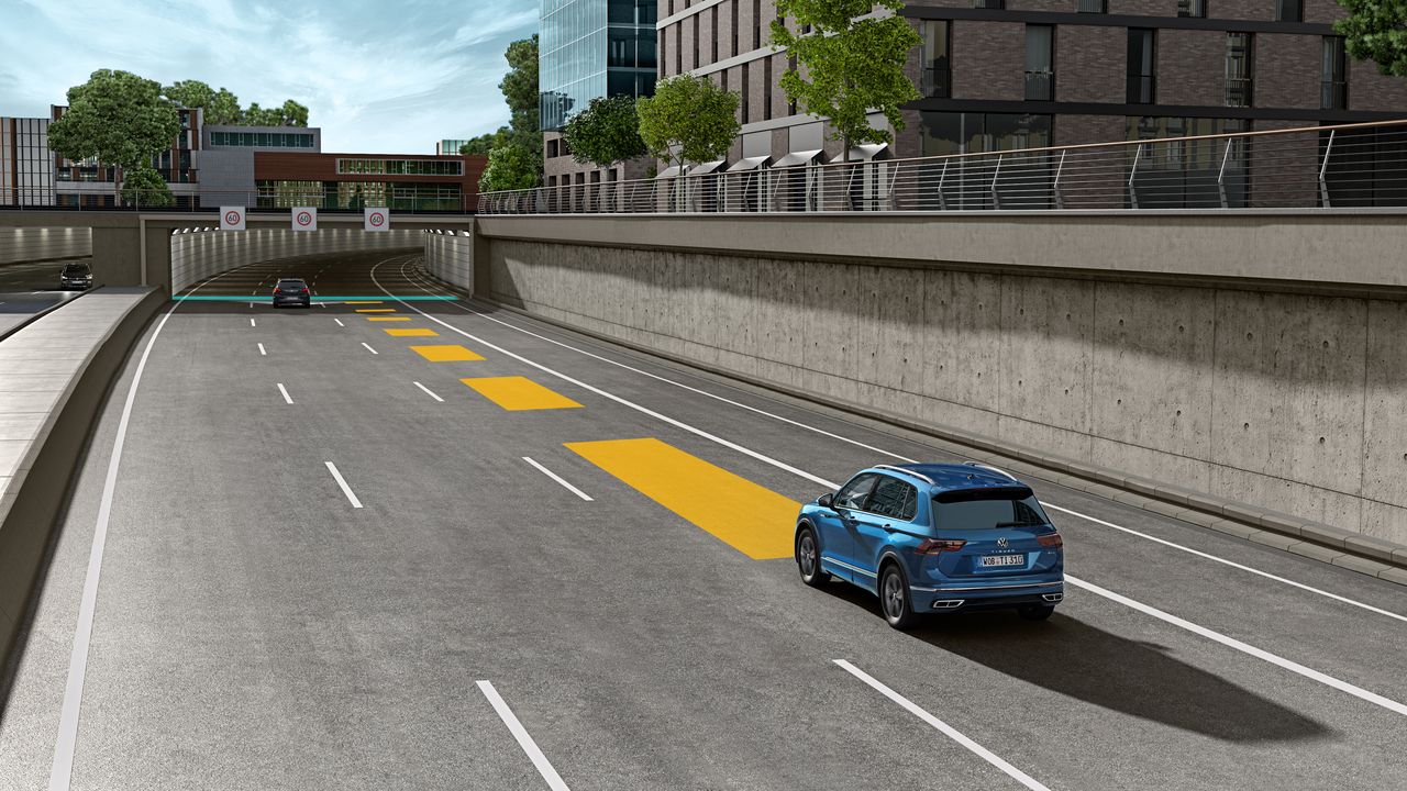 Automobil jazdí po ceste smerom do tunela v mestskej oblasti. Na ceste sú vyznačené žlté pruhy , ktoré znázorňujú adaptívny tempomat ACC. V pozadí sú moderné budovy a zeleň.