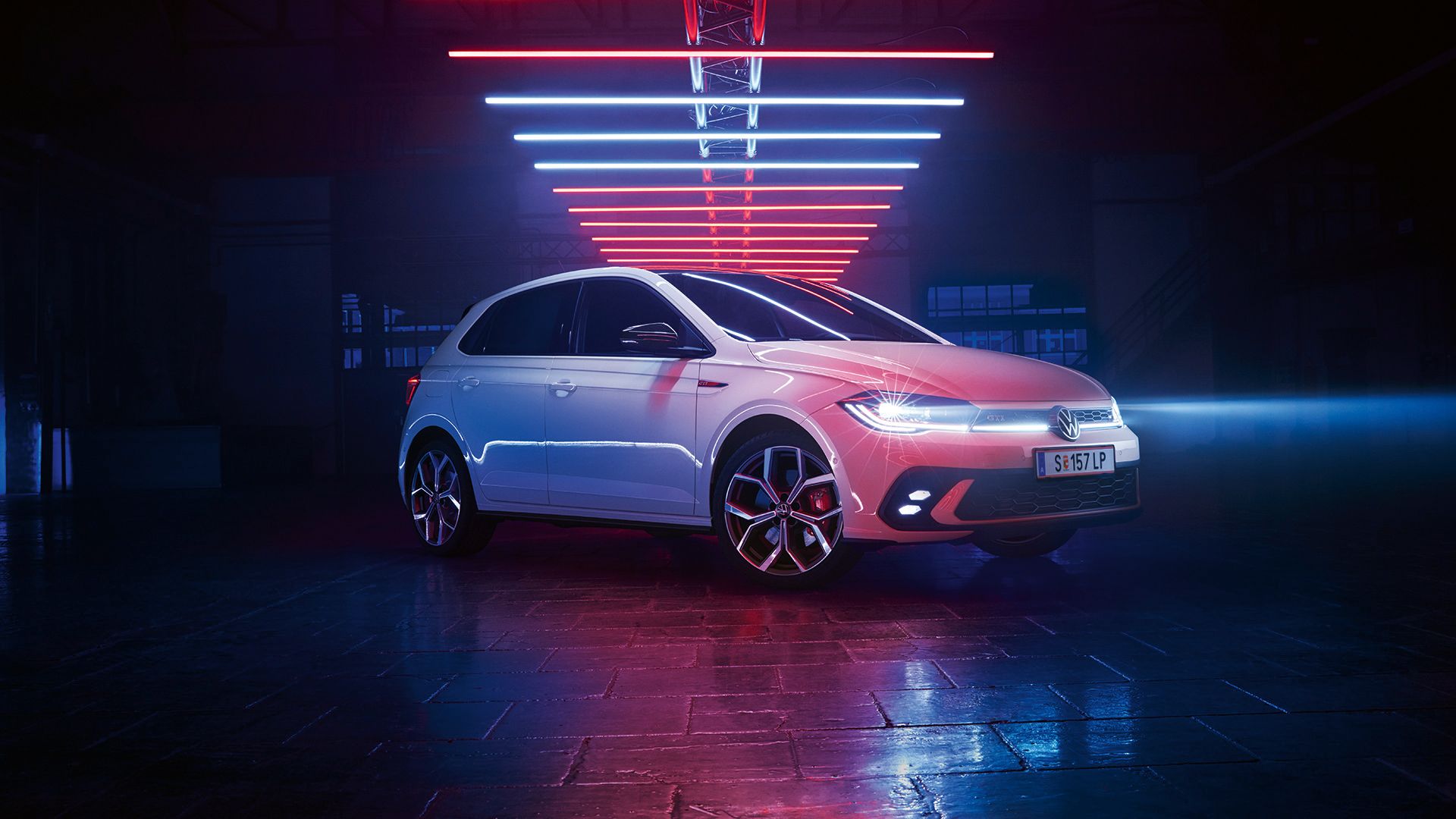 Aufnahme eines weißen VW Polo GTI, der mit angeschalteten LED-Scheinwerfern in einer dunklen Halle unter Leuchtelementen steht.