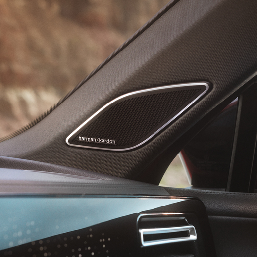 Detailný záber na reproduktor značky Harman Kardon, ktorý je zabudovaný v A-stĺpiku automobilu. Na pozadí je rozmazaný výhľad na vonkajšie prostredie.