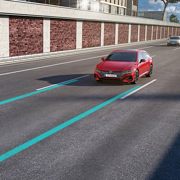 Lane assist a Side assist zobrazené ako dve modré línie jedného jazdného pruhu, v ktorom jazdí červené vozidlo. Vo vedľajšom pruhu ide druhé strieborné vozidlo. V pozadí vidieť oporný múr a modernú stavbu a stromy.