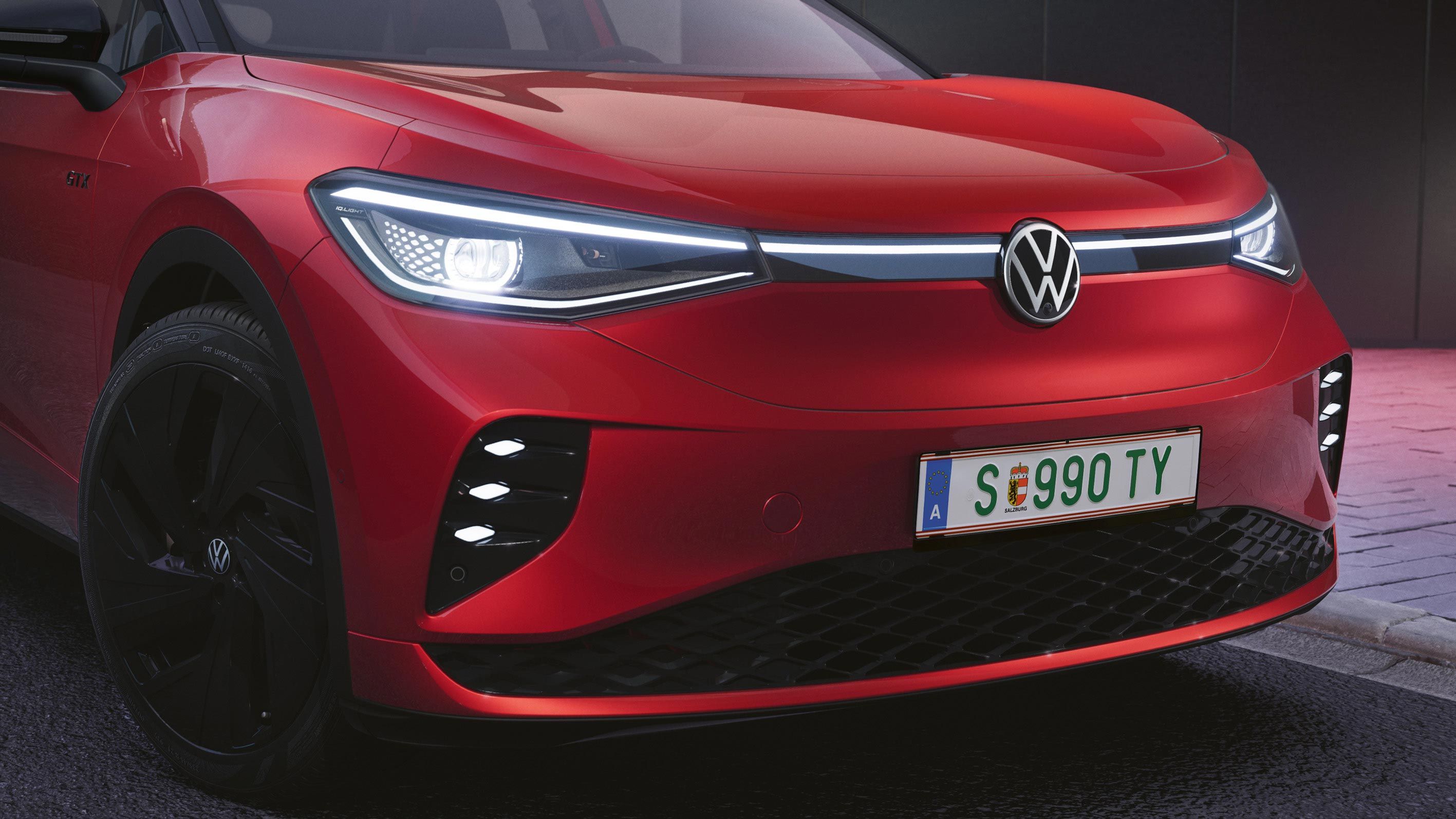 VW ID.4 GTX bočný pohľad na predné partie. Vozidlo má zapnuté moderné predné svetlomety. Červený lak vozidla dopĺňajú čierne disky kolies a čierny kryty vonkajších spätných zrkadiel.