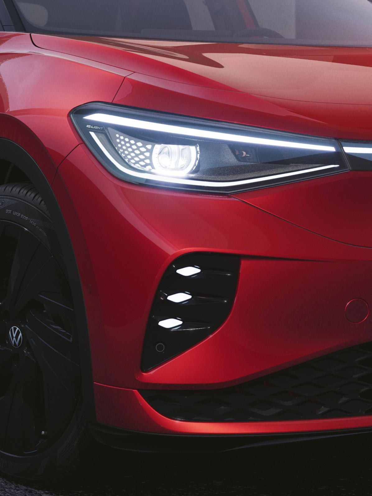 VW ID.4 GTX bočný pohľad na predné partie. Vozidlo má zapnuté moderné predné svetlomety. Červený lak vozidla dopĺňajú čierne disky kolies a čierny kryty vonkajších spätných zrkadiel.