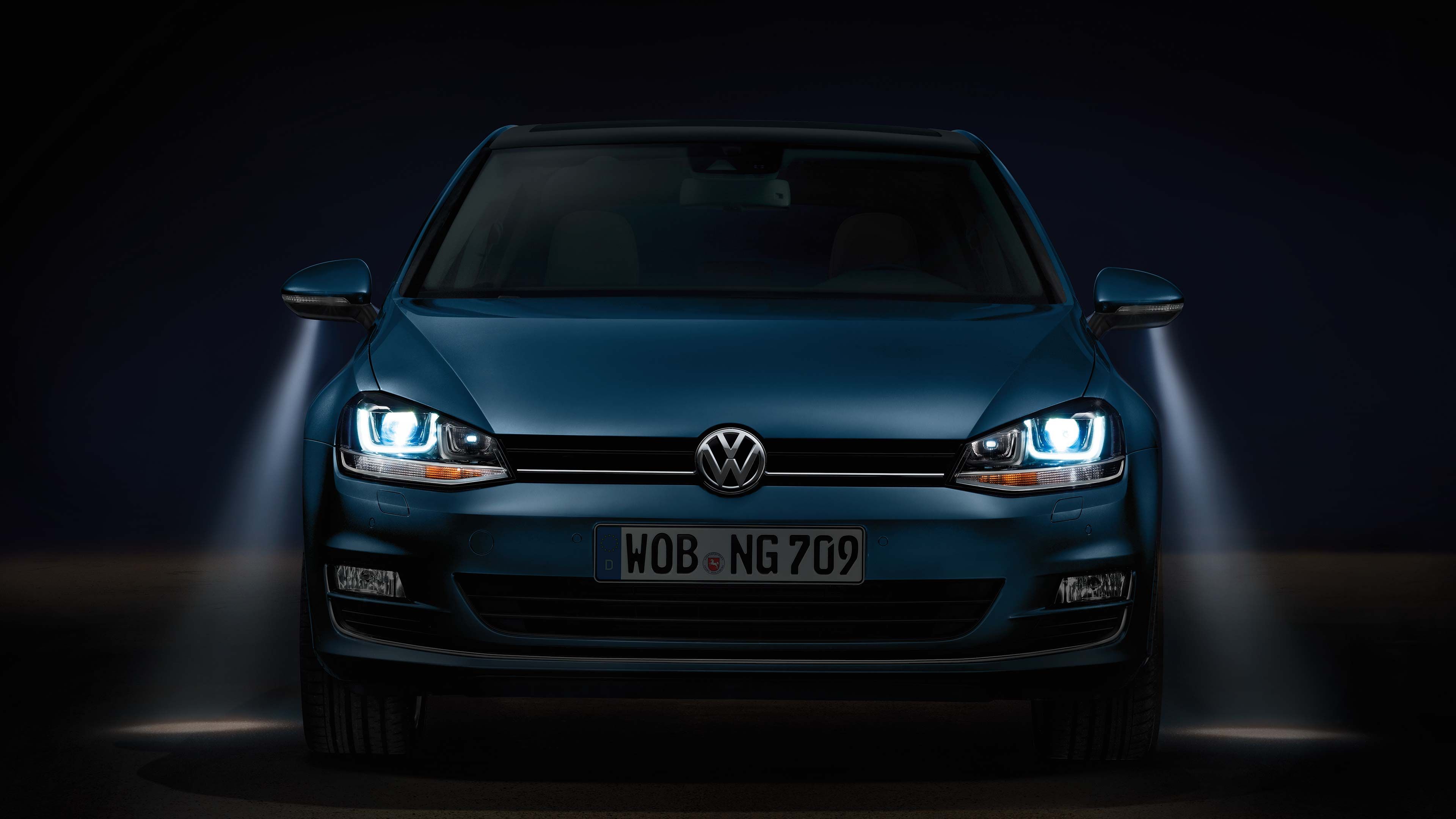 Tmavomodré auto značky Volkswagen so zapnutými svetlami vrátane osvetlenia okolia vozidla pri vystupovaní v tmavom prostredí. Špz nesie nápis WOB - NG 709. Predná maska auta je dobre viditeľná.