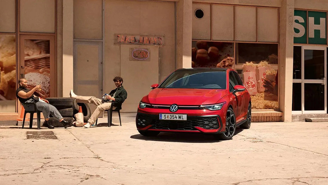 Pohľad spredu na VW Golf GTI, ktorý je zaparkovaný pred staršou budovou. Vedľa auta sedia dvaja ľudia. V budove je obchod.