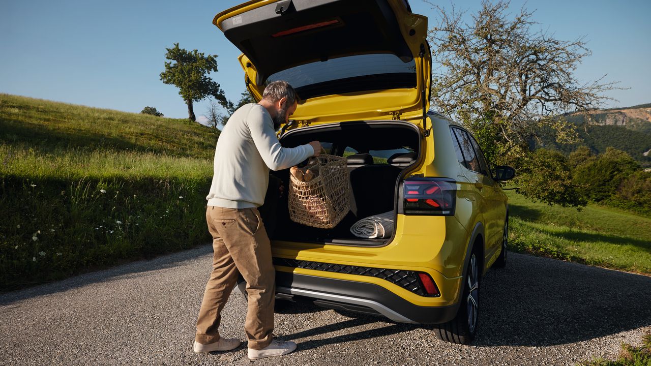 Muž vloží veľký piknikový kôš do batožinového priestoru žltého VW T-Cross, ktorý je zaparkovaný na ceste v hornatej krajine.