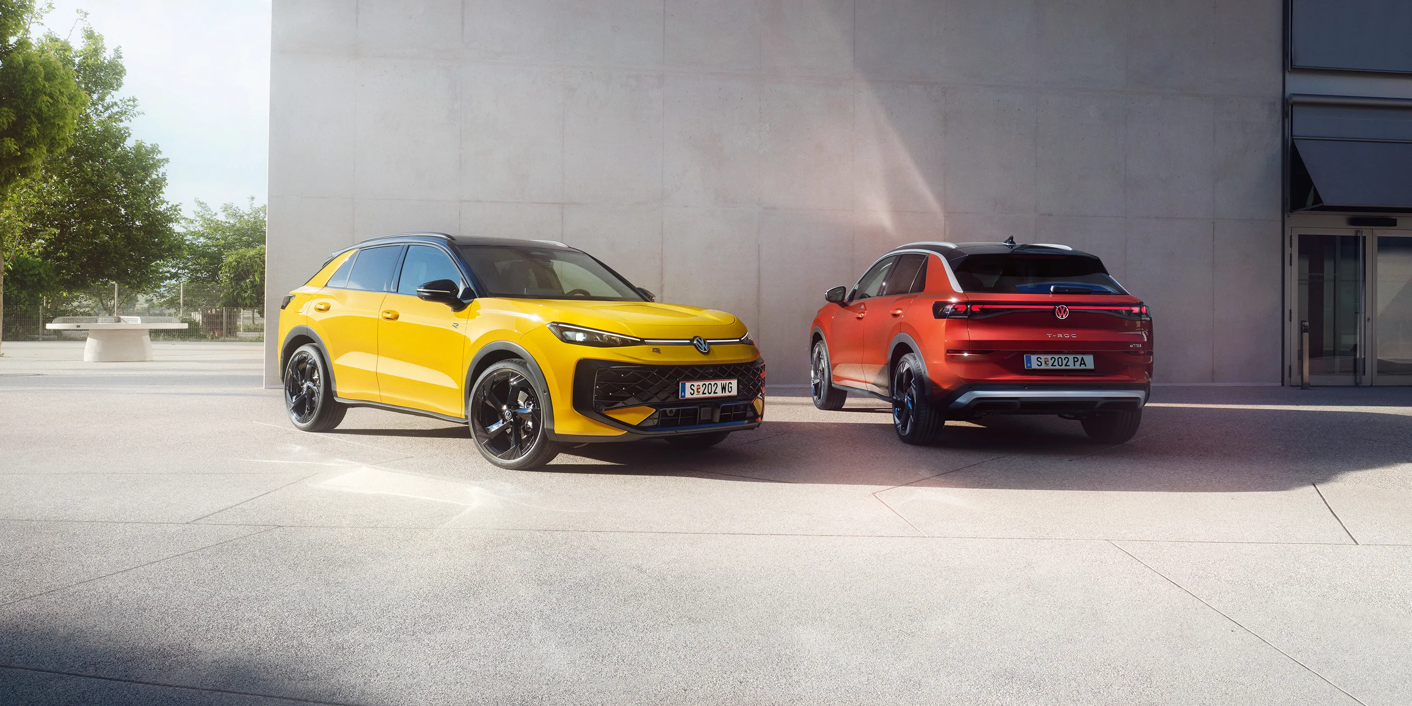 Žltý VW T-Roc R-Line a červený VW T-Roc Style stoja vedľa seba pred modernou budovou.
