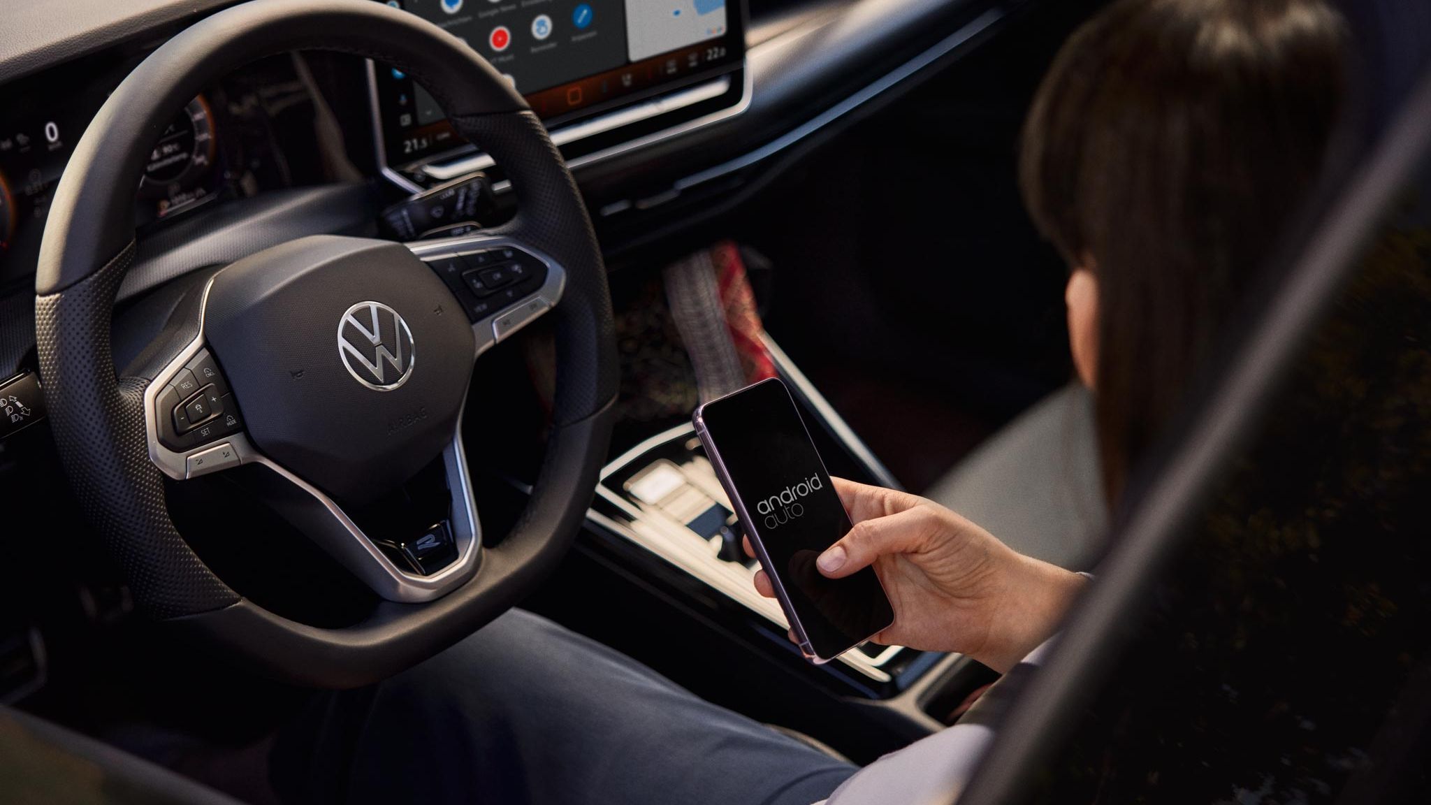 Žena sedí vo vozidle Volkswagen a drží telefón so systémom Android Auto. Na volante je logo Volkswagen a v pozadí vidieť palubnú dosku auta s veľkým displejom systému infotainment.