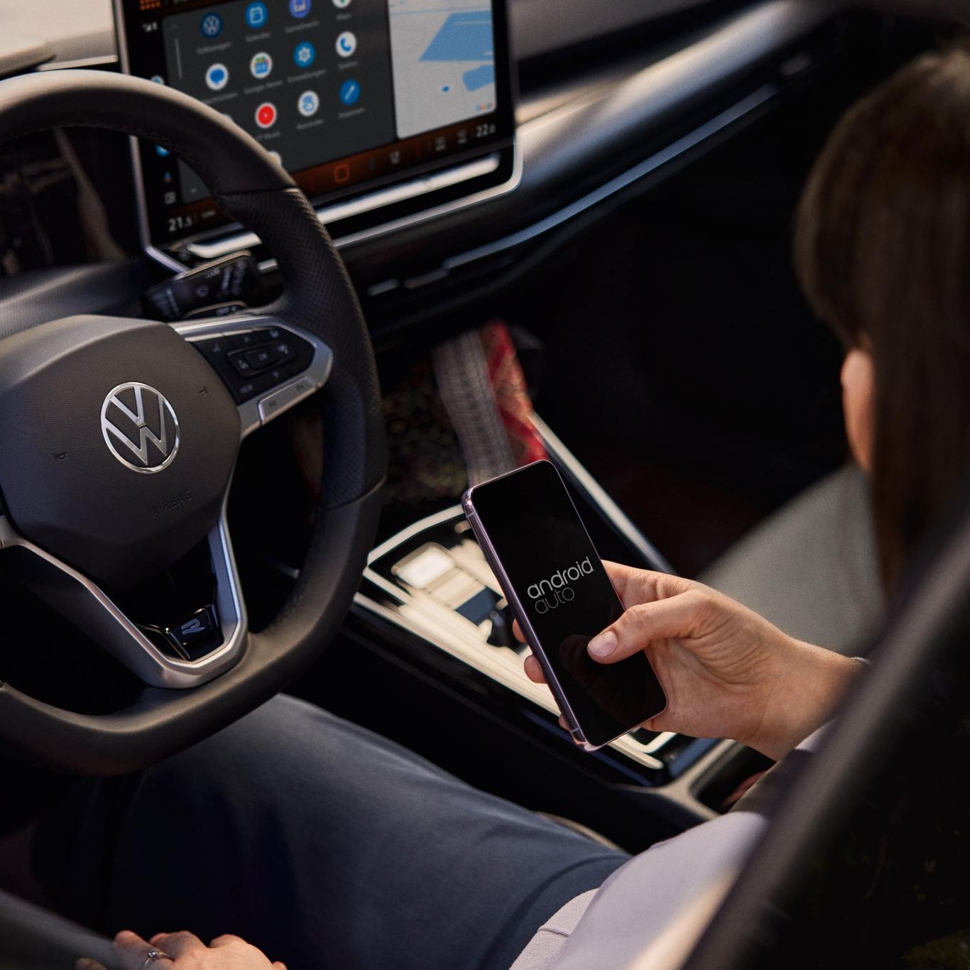 Žena sedí vo vozidle Volkswagen a drží telefón so systémom Android Auto. Na volante je logo Volkswagen a v pozadí vidieť palubnú dosku auta s veľkým displejom systému infotainment.