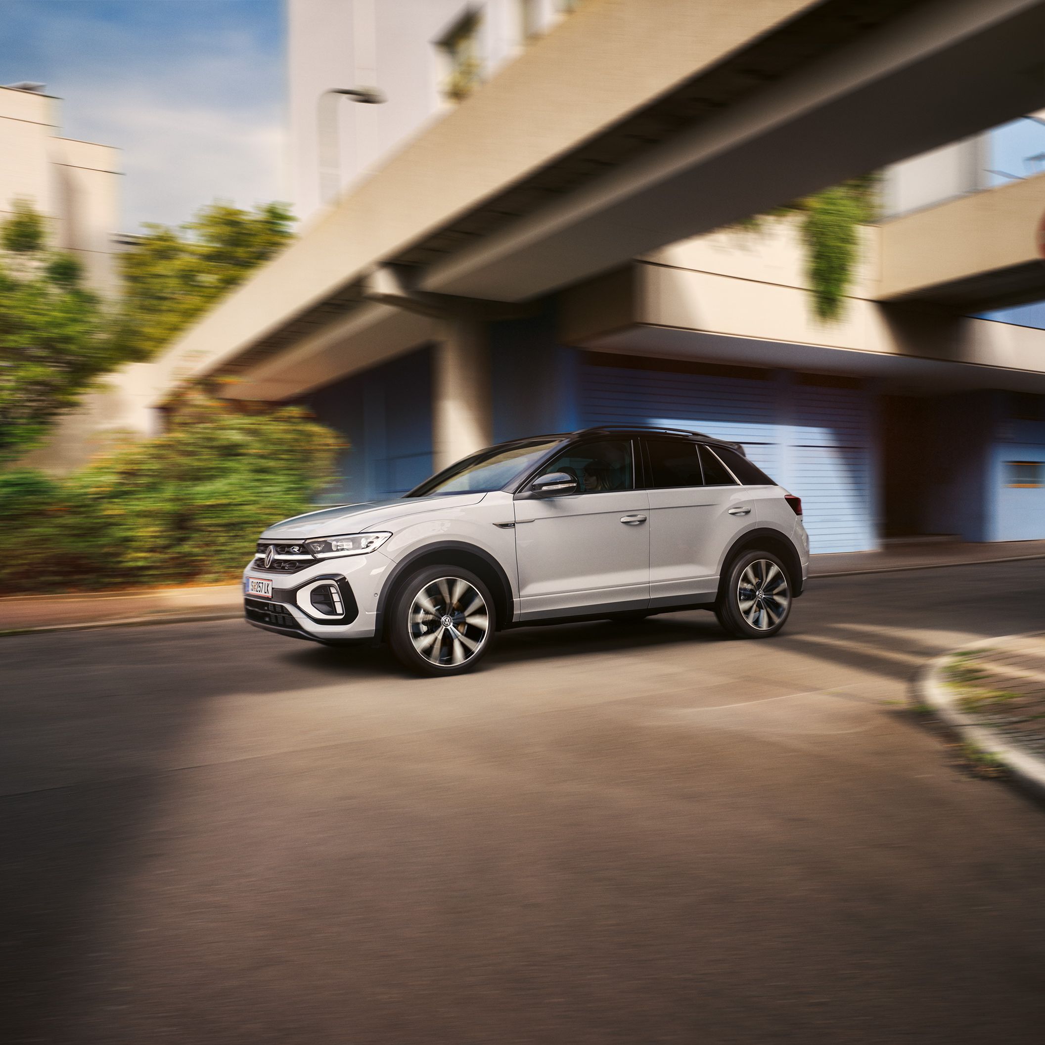 Strieborné SUV jazdí po ulici v meste. Pozadie tvorí moderná architektúra a zeleň. 