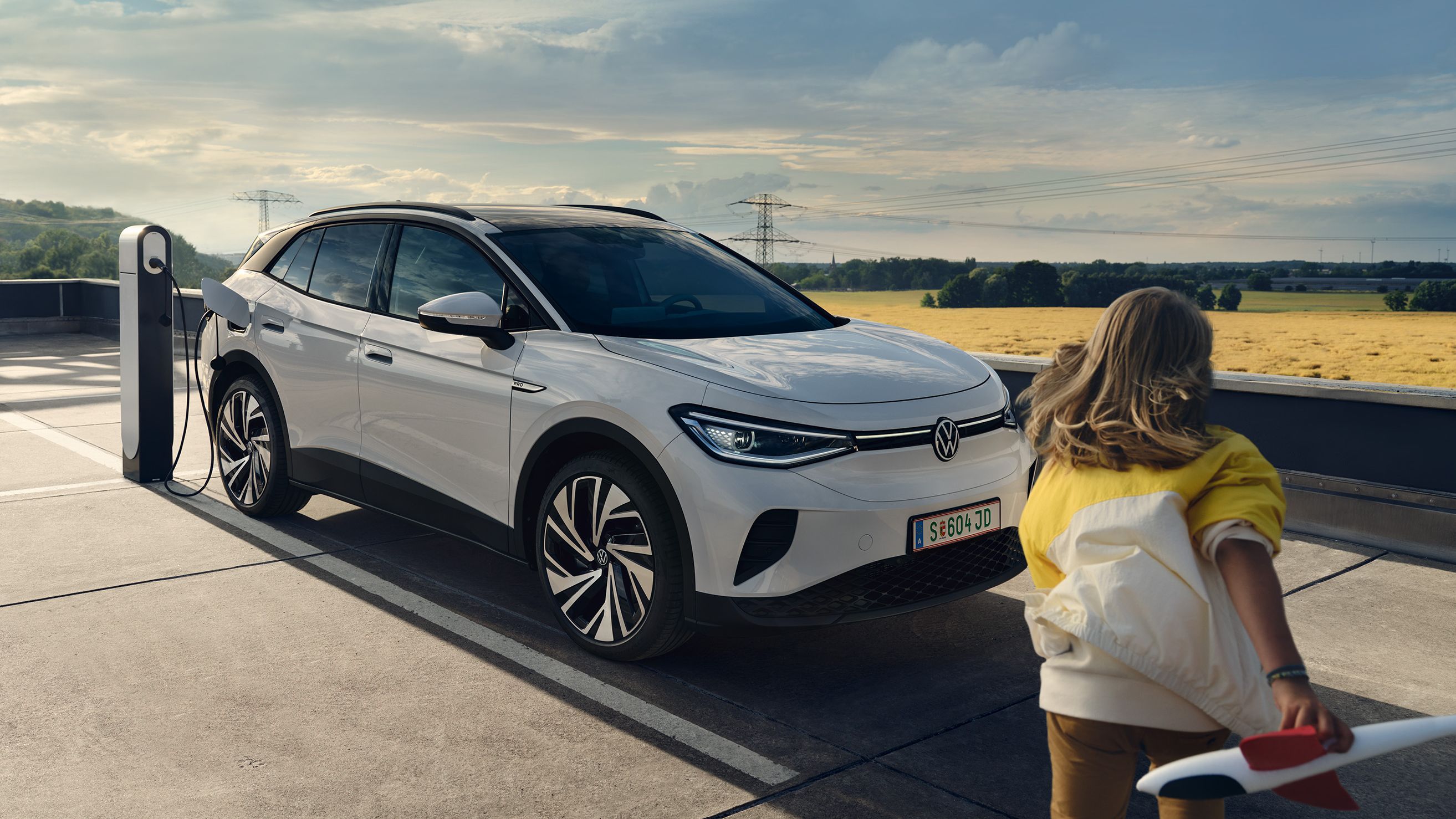 Žena drží model lietadla a prichádza k bielemu elektrickému SUV, ktoré sa nabíja na nabíjacej stanici pod modrou oblohou. Auto je zaparkované na vonkajšom parkovisku. V pozadí sa rozprestiera lúka a niekoľko stromov.