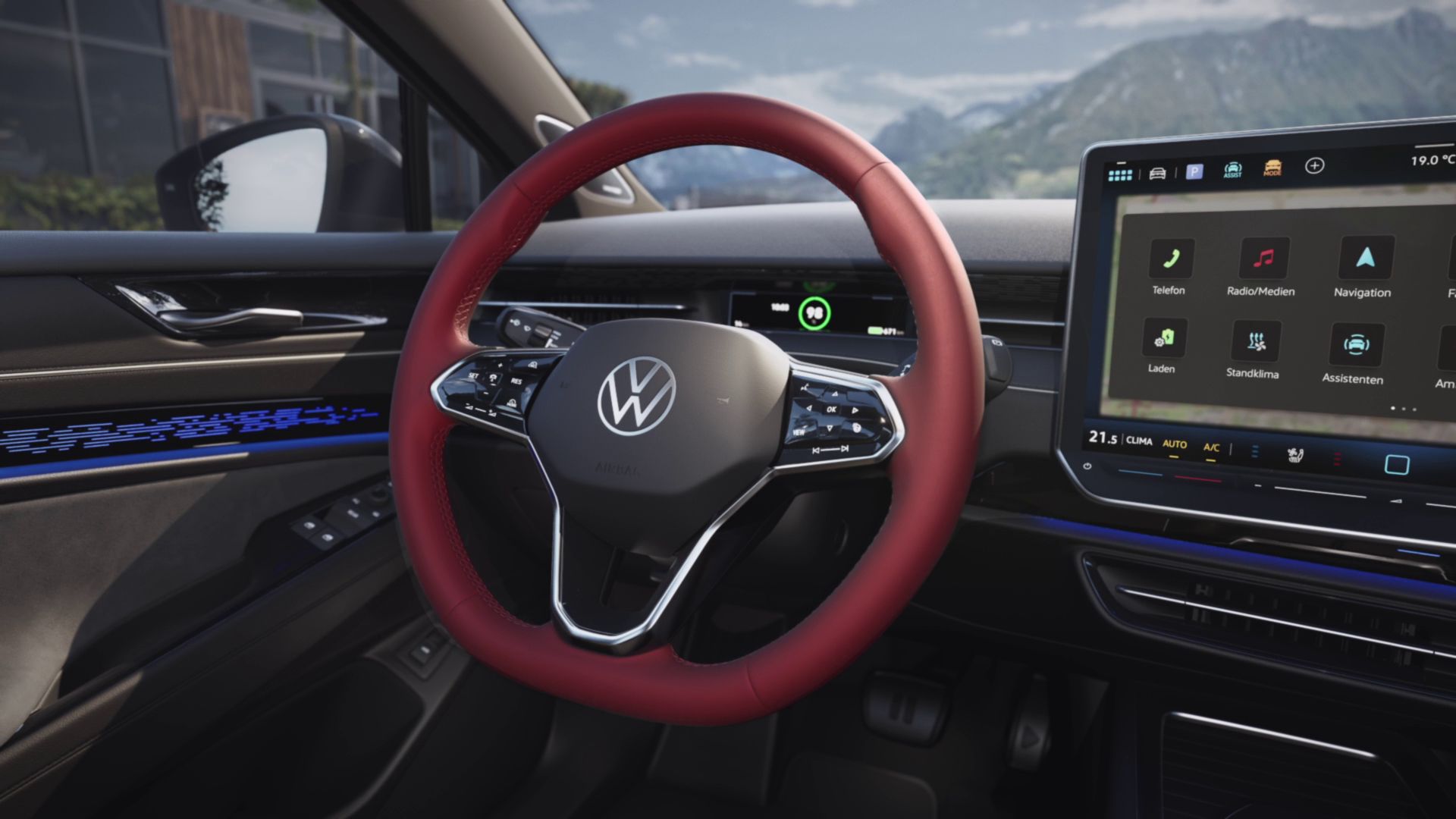 Na obrázku je interiér moderného auta. V popredí je vidieť volant s červeným poťahom a logom VW, vpravo od neho je veľký displej s rôznymi položkami menu. V pozadí sú viditeľné hory a cesta.