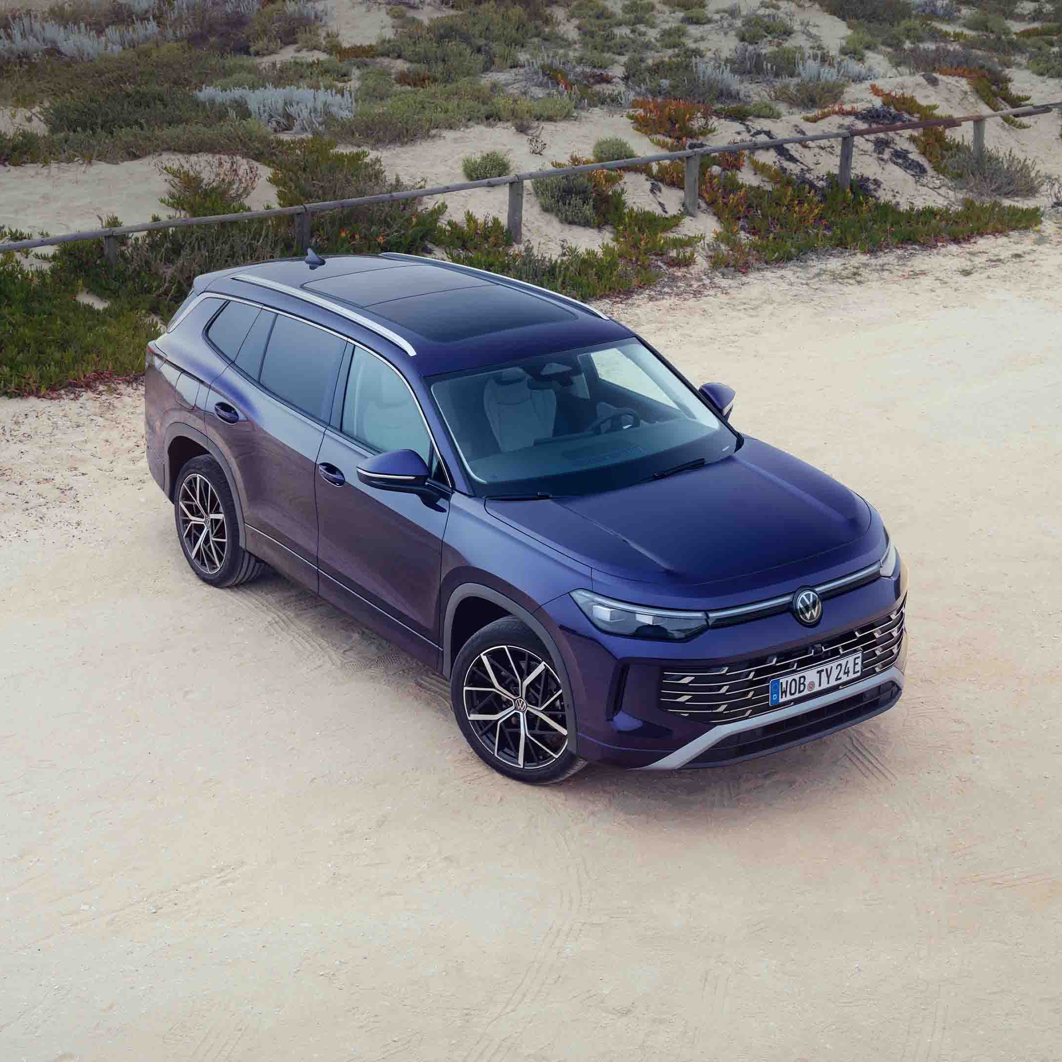 VW Tayron zaparkovaný na voľnom priestranstve pri piesočnej pláži. Okolo sú rastliny a drevený plot. Automobil má moderný dizajn.