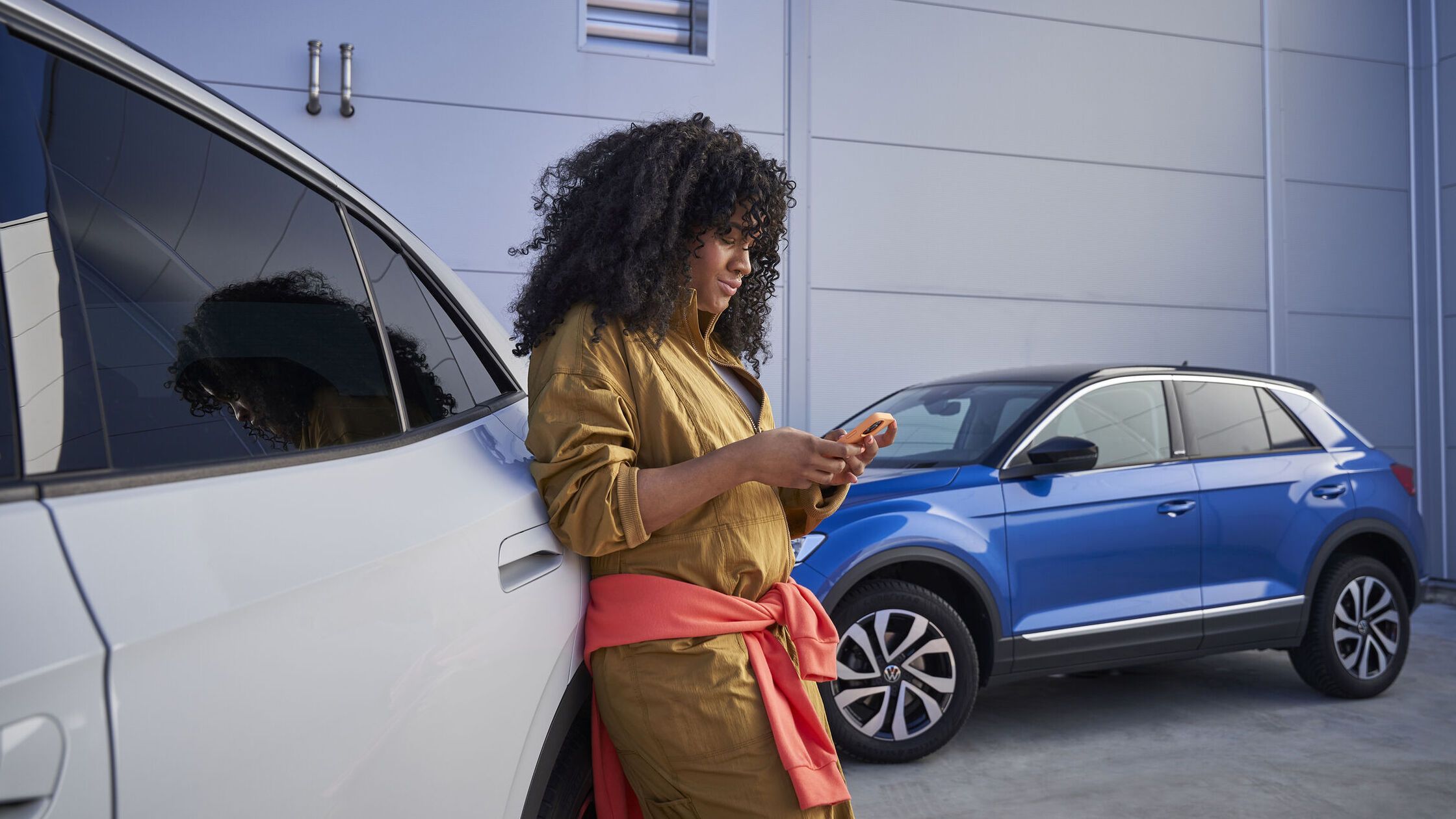 Kučeravá žena sa opiera o biele vozidlo a drží mobilný telefón. Vedľa nej je zaparkované modré vozidlo Volkswagen.