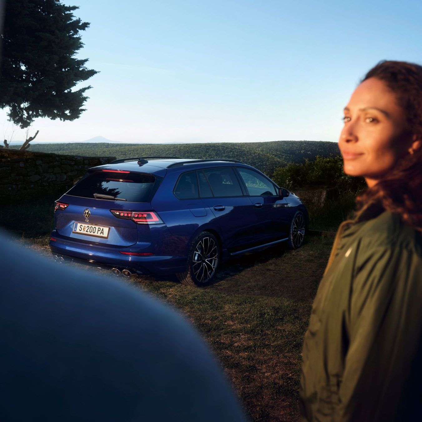 Modrý VW Golf R Variant zaparkovaný na trávniku v prírode. V popredí stoja dvaja ľudia. Muž je otočený chrbtom a žena sa pozerá na muža.