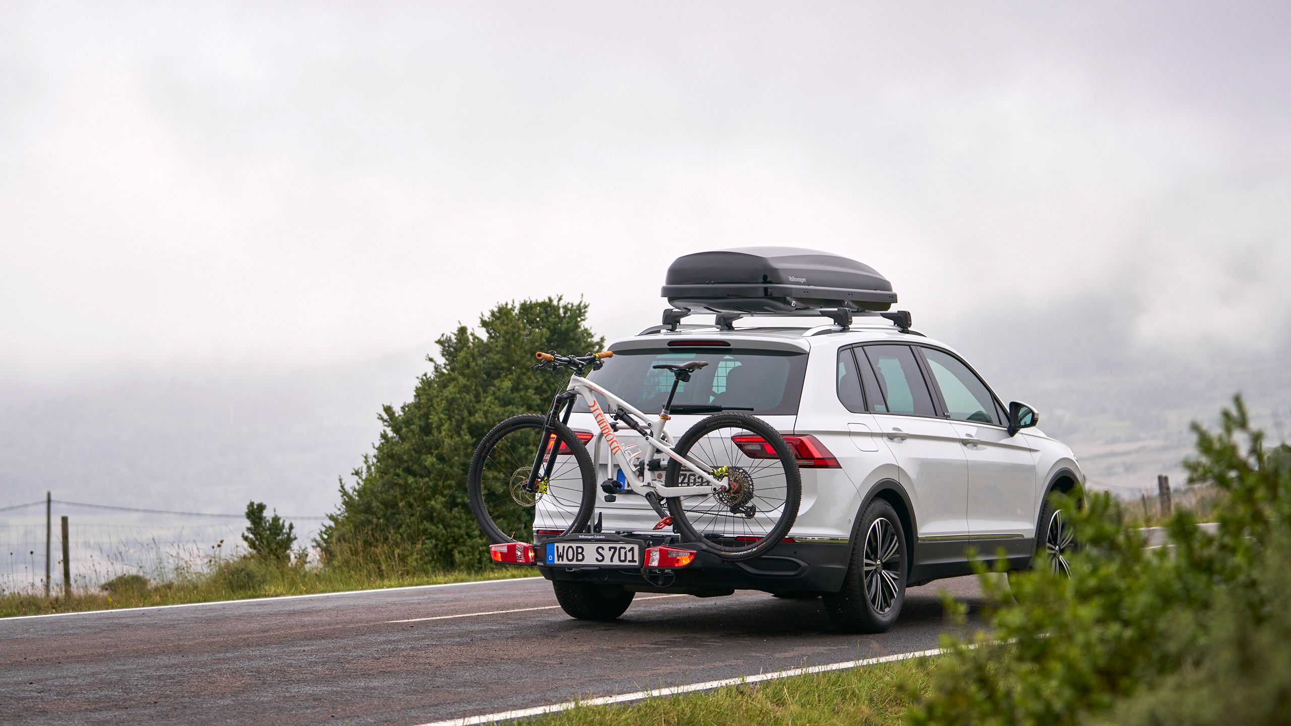 VW Tiguan so strešným boxom a nosičom bicyklov z originálneho príslušenstva Volkswagen vrátane bicykla na poľnej ceste
