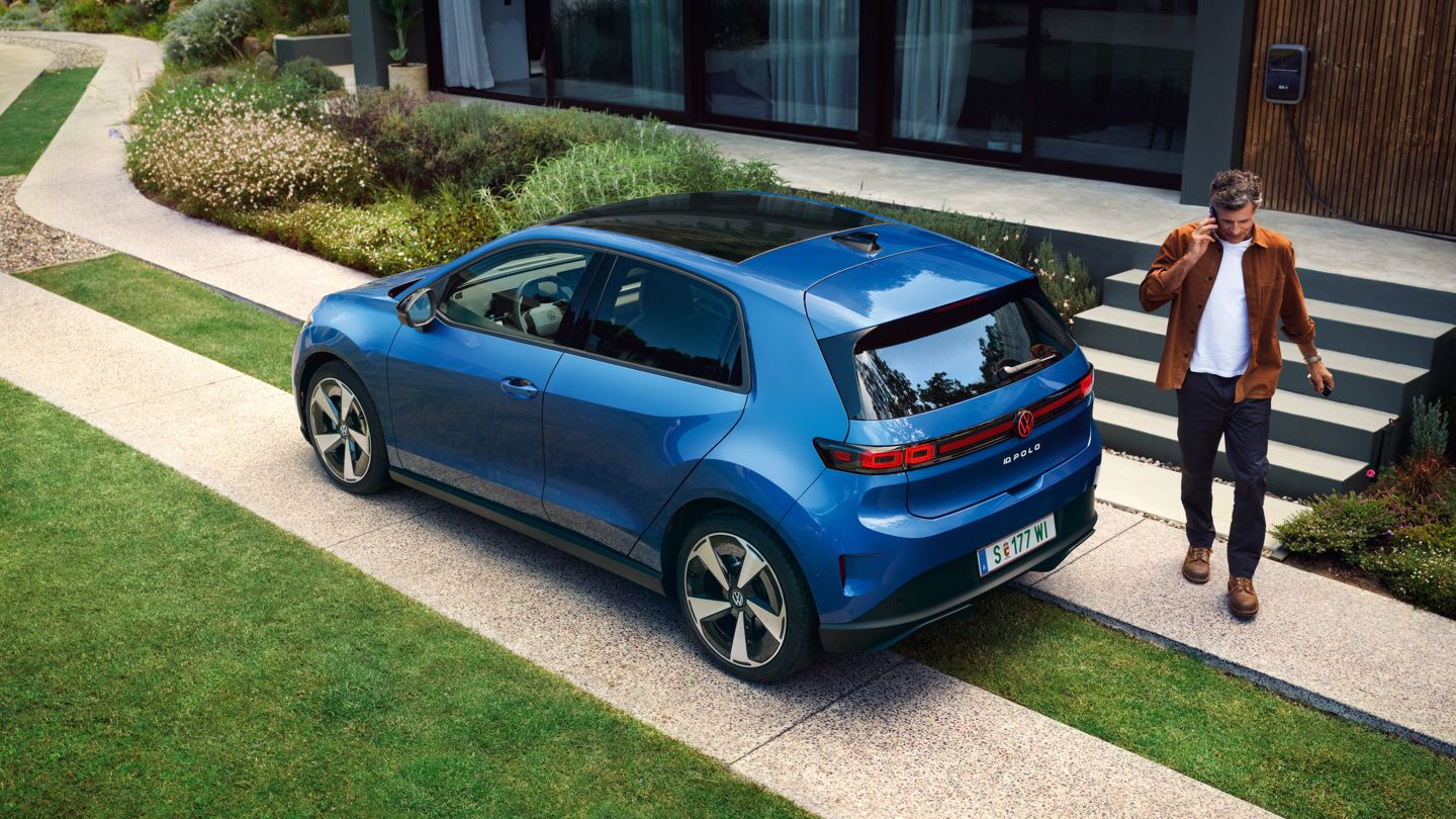 Modrý VW ID. Polo Style stojí zaparkovaný pred obytným domom so zeleňou, za vozidlom prechádza muž.